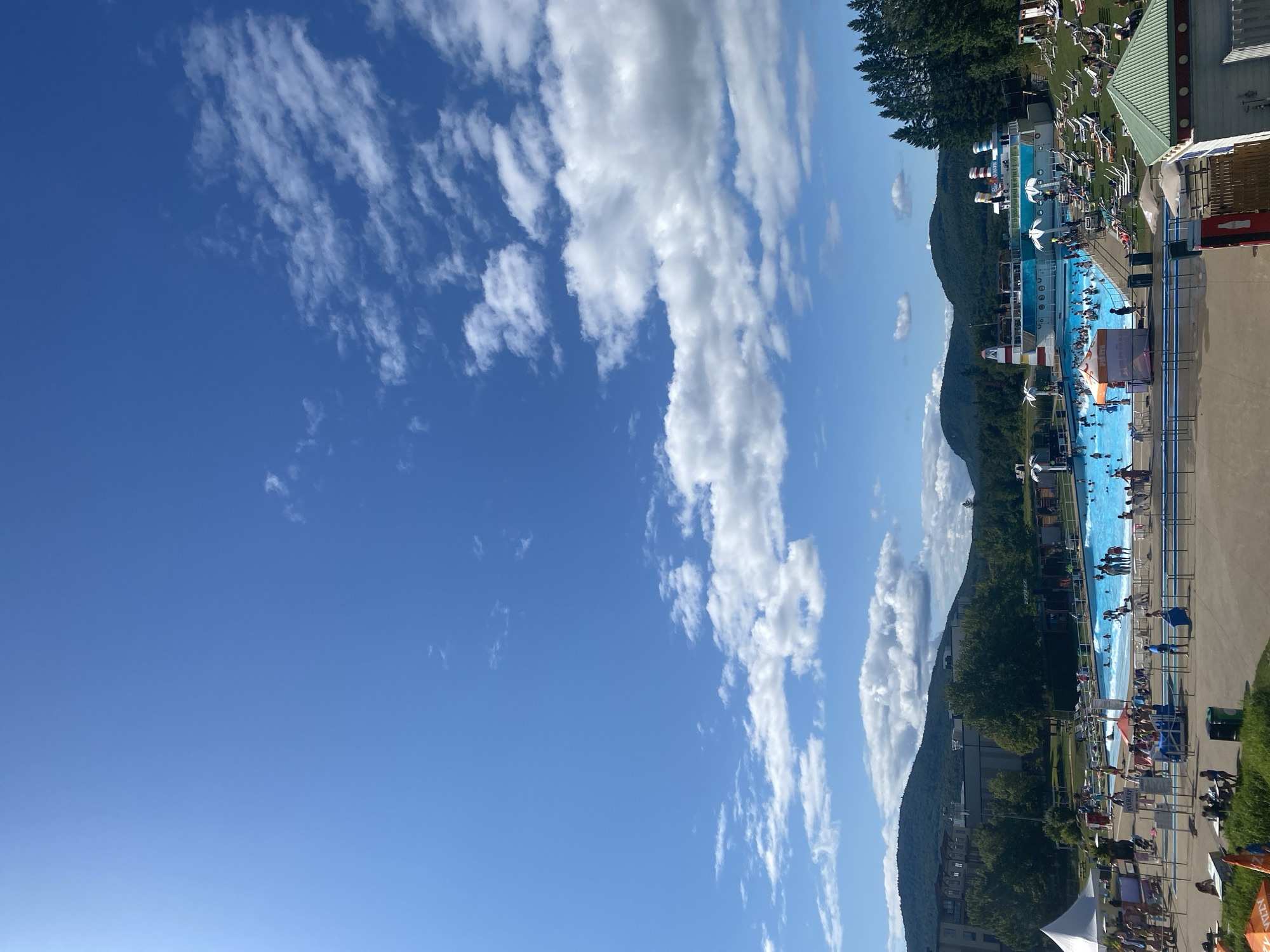 This is a daytime, outdoor photograph of a water park, likely at Montmorency Falls, in Saint-Gabriel-de-Valcartier, Canada. The sky is a vibrant blue with scattered white clouds. In the foreground, there is a large, blue wave pool filled with numerous people swimming and wading. Surrounding the pool are lounge chairs with people relaxing on them, as well as food and drink stands. A large, ship-like structure with slides and water features is positioned at one end of the pool. Beyond the water park, a wooded area and rolling hills are visible, with buildings and a ski lift structure in the distance. The overall atmosphere appears to be one of leisure and recreation.