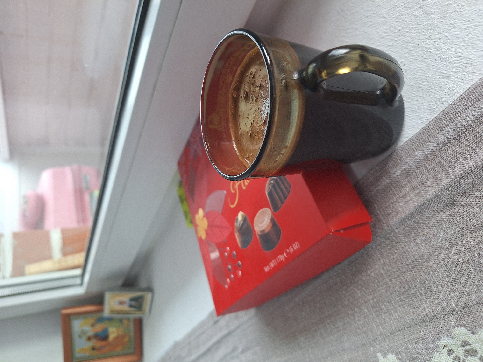 A close-up, eye-level shot shows a dark brown mug filled with black coffee and a foam top, positioned on a white windowsill next to a red box of chocolates. The mug has a dark, translucent handle and is slightly to the right of the center of the frame. To the left of the mug is a red box with gold lettering and floral designs, containing several chocolates visible on its surface. Behind the chocolates, two small religious icons are placed against the white windowsill. The background beyond the windowsill is blurred but suggests an indoor environment with a window offering a view of possibly outdoor structures. The overall scene evokes a quiet, contemplative moment. The presence of religious icons suggests a possible domestic setting in Vologda, Russia, where such items are common.