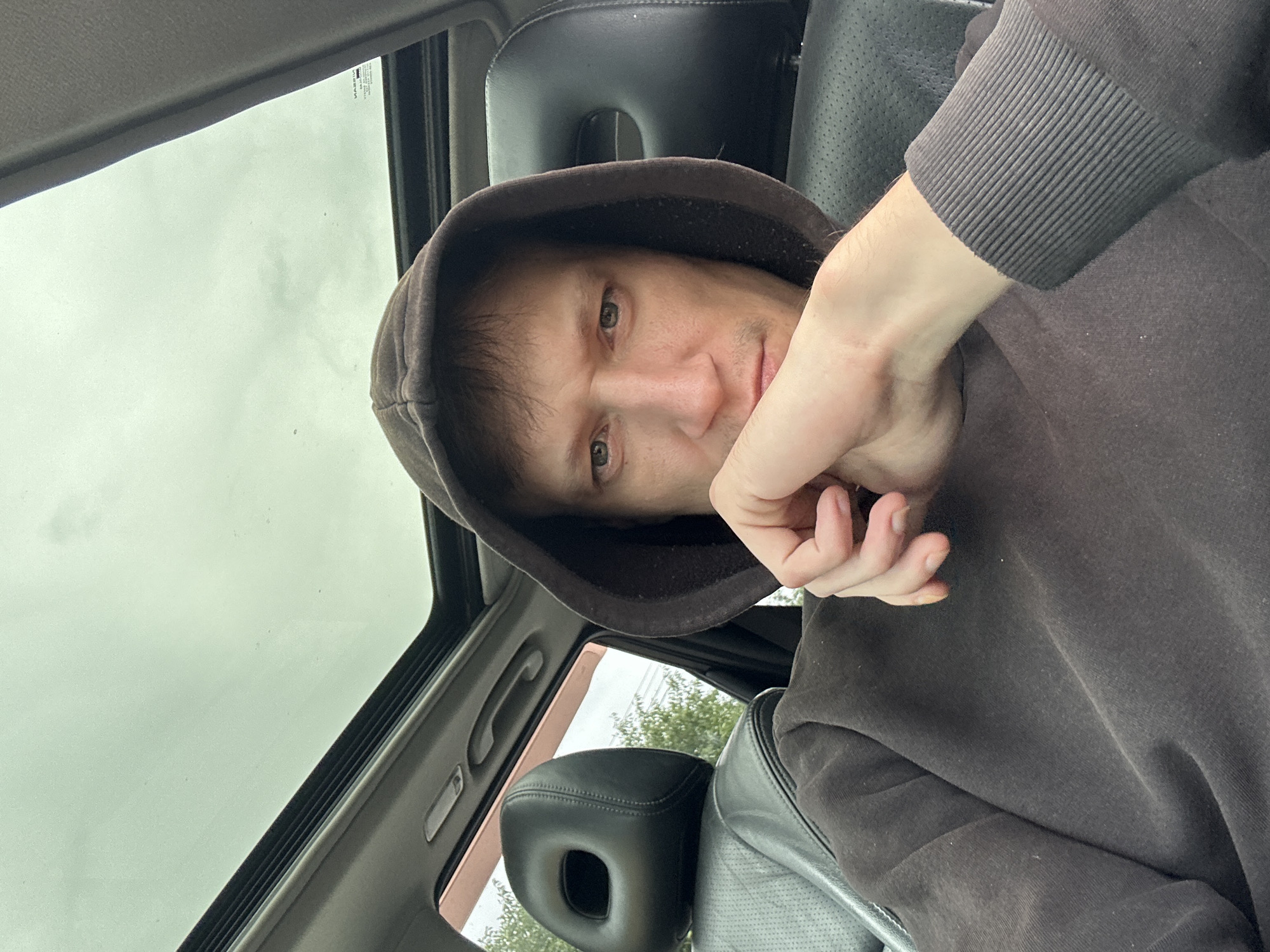 A man is inside a car, with a sunroof open above him. He is wearing a dark brown hooded sweatshirt and his face is partially obscured by his hand, which is resting on his chin. The interior of the car is visible, including a black leather headrest. Outside the car, trees and sky are visible. The image was taken on July 23, 2023, at 10:47 AM, at coordinates 49.3919° N, 30.0611° E, indicating the location is in the vicinity of Makiv, Ukraine.