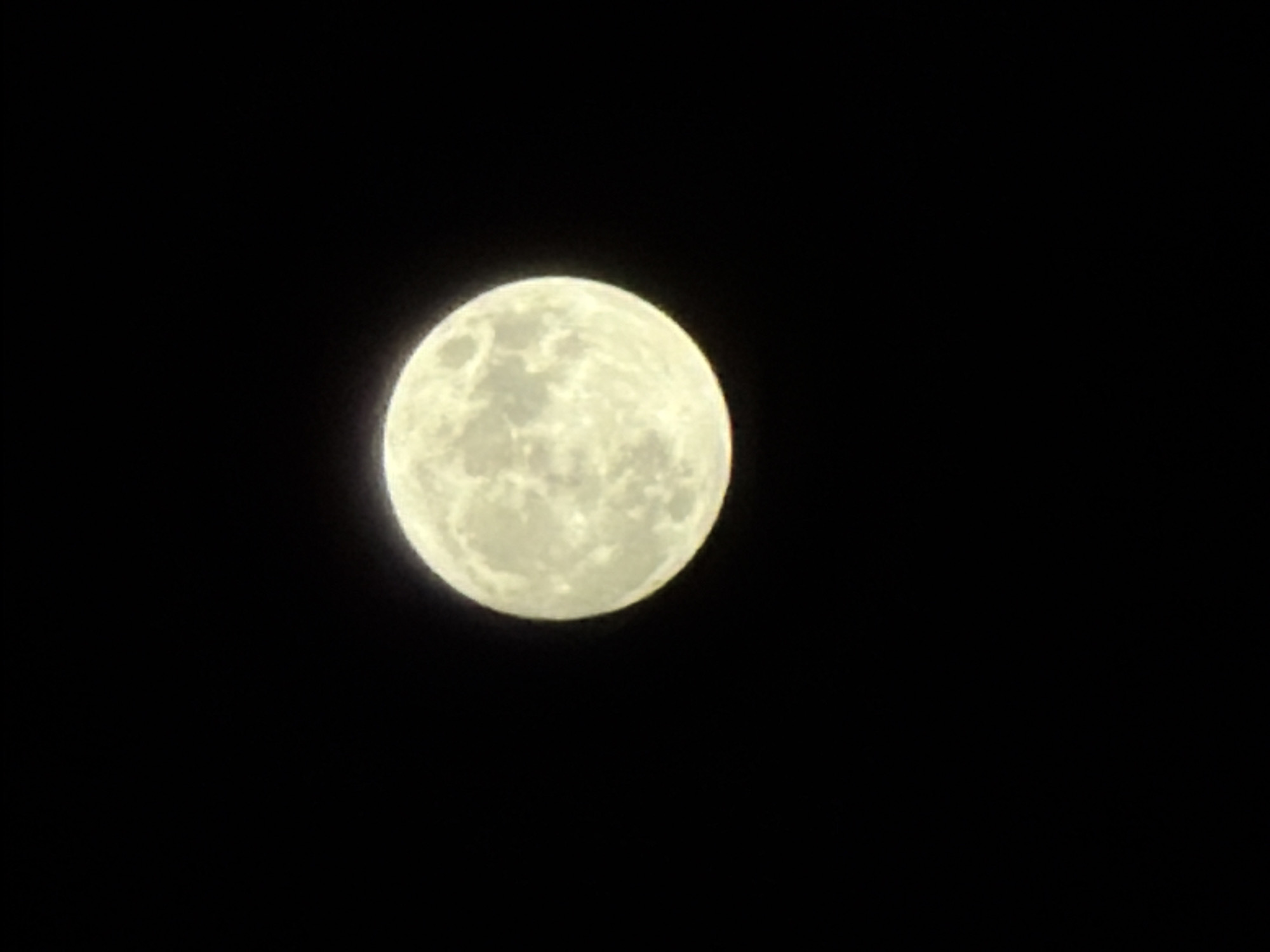 The image captures a radiant full moon set against a dark, clear night sky. The moon is the sole dominant subject, appearing as a luminous sphere with visible topographical features, including darker mare regions and lighter, cratered areas, giving it a textured, ethereal glow. The celestial body is positioned slightly off-center in the frame, illuminating the void around it. The background is an unblemished expanse of deep black, indicating a cloudless night devoid of other stars or atmospheric phenomena. There are no people, man-made structures, or other natural objects discernible in the frame, highlighting the moon in isolation. The scene depicts no specific activity or event, merely a static view of the moon at nighttime. The provided context indicates the image was taken in Izhevsk, Russia. No visible text is present within the media file.