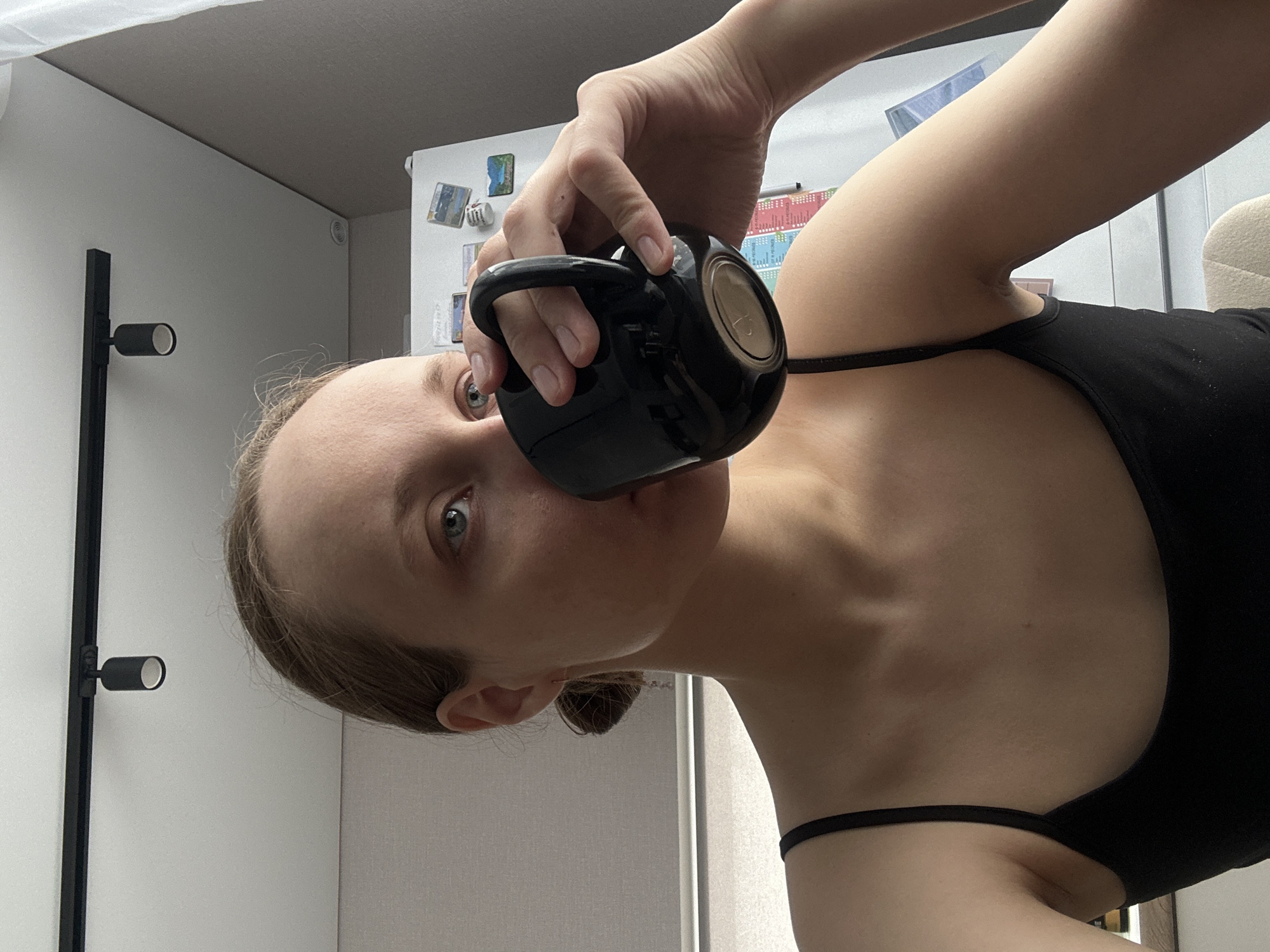 A woman is holding a black mug to her lips, seemingly drinking from it. She is wearing a black tank top. The background includes a light-colored wall with a black, minimalist lamp fixture, and a refrigerator adorned with magnets. There is no indication of the location being Подшивалово, Russia.