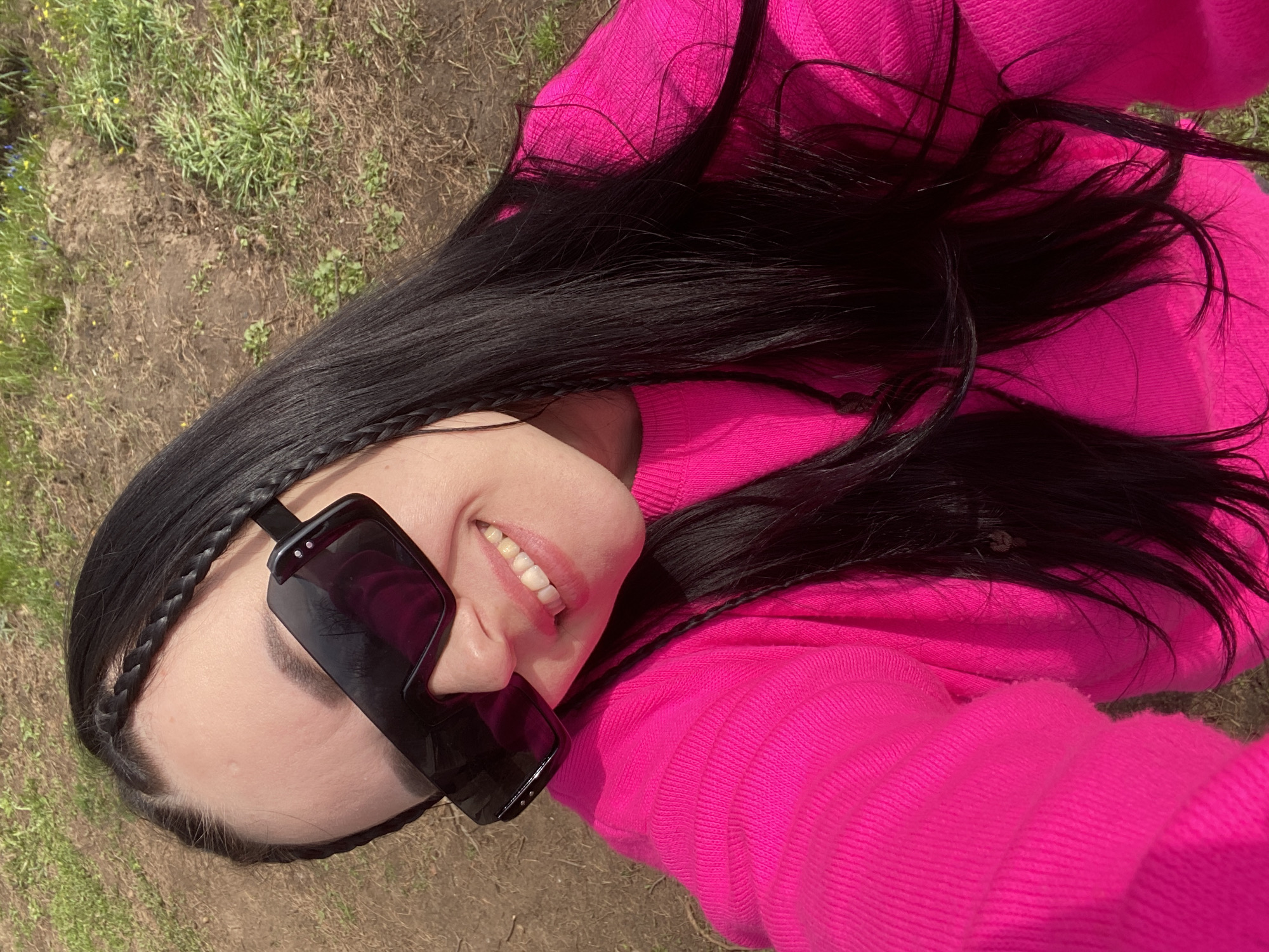 A high-angle selfie captures a person smiling broadly outdoors in Chaikovsky, Russia, under bright daylight. The subject, seen from the shoulders up, is looking upwards at the camera with a joyful expression, revealing her teeth. She wears prominent dark, shield-style sunglasses that cover a portion of her face. Her long, straight, dark hair is styled with a small braid near her forehead, and loose strands frame her face. She is dressed in a vibrant, hot pink, ribbed knit sweater or top. The background shows an outdoor environment with patches of green grass and areas of exposed brown soil, typical of a natural, unpaved setting. The image conveys a cheerful and relaxed mood. No text is visible in the media file.
