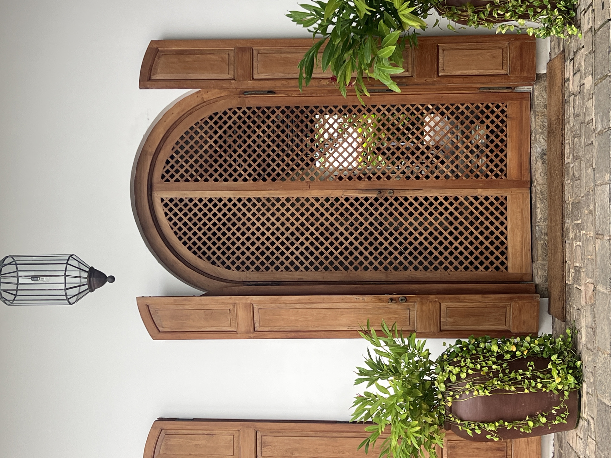 An outdoor scene features a weathered wooden arched door with a lattice pattern, flanked by open wooden shutters. Two large, dark brown planters with lush green trailing plants are positioned on either side of the doorway, resting on a cobblestone pathway. A dark-colored doormat lies in front of the door. Above the door, a decorative cage-like lantern hangs from a white wall. The architecture suggests a colonial style, consistent with the historic Fort area of Galle, Sri Lanka, known for its Dutch colonial buildings. No people are present in the frame.