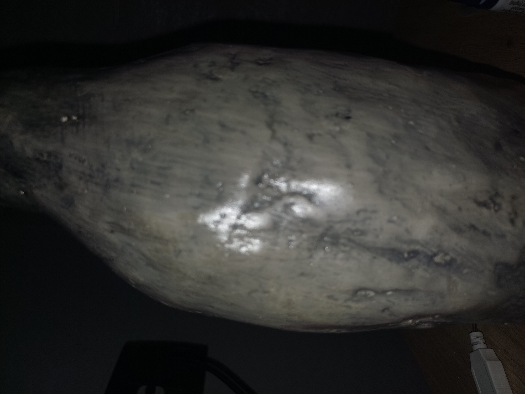 A close-up, low-light, slightly out-of-focus shot captures the textured surface of a dark grey, potentially ceramic or stone, vase or bottle. The object is predominantly a mottled grey with darker, irregular patches and lighter, almost reflective areas, suggesting a glossy or wet surface. Subtle ridges and indentations are visible, indicating an uneven, possibly handmade, texture. To the left of the object, a black electronic device with a curved cable is partially visible, and below it, a white USB connector with a black cable is on a wooden surface. The background is dark, obscuring much of the environment, but a hint of a dark vertical surface is visible to the far right. There are no people present in the image. The scene does not provide any specific information to indicate its location in Sulymivka, Ukraine.