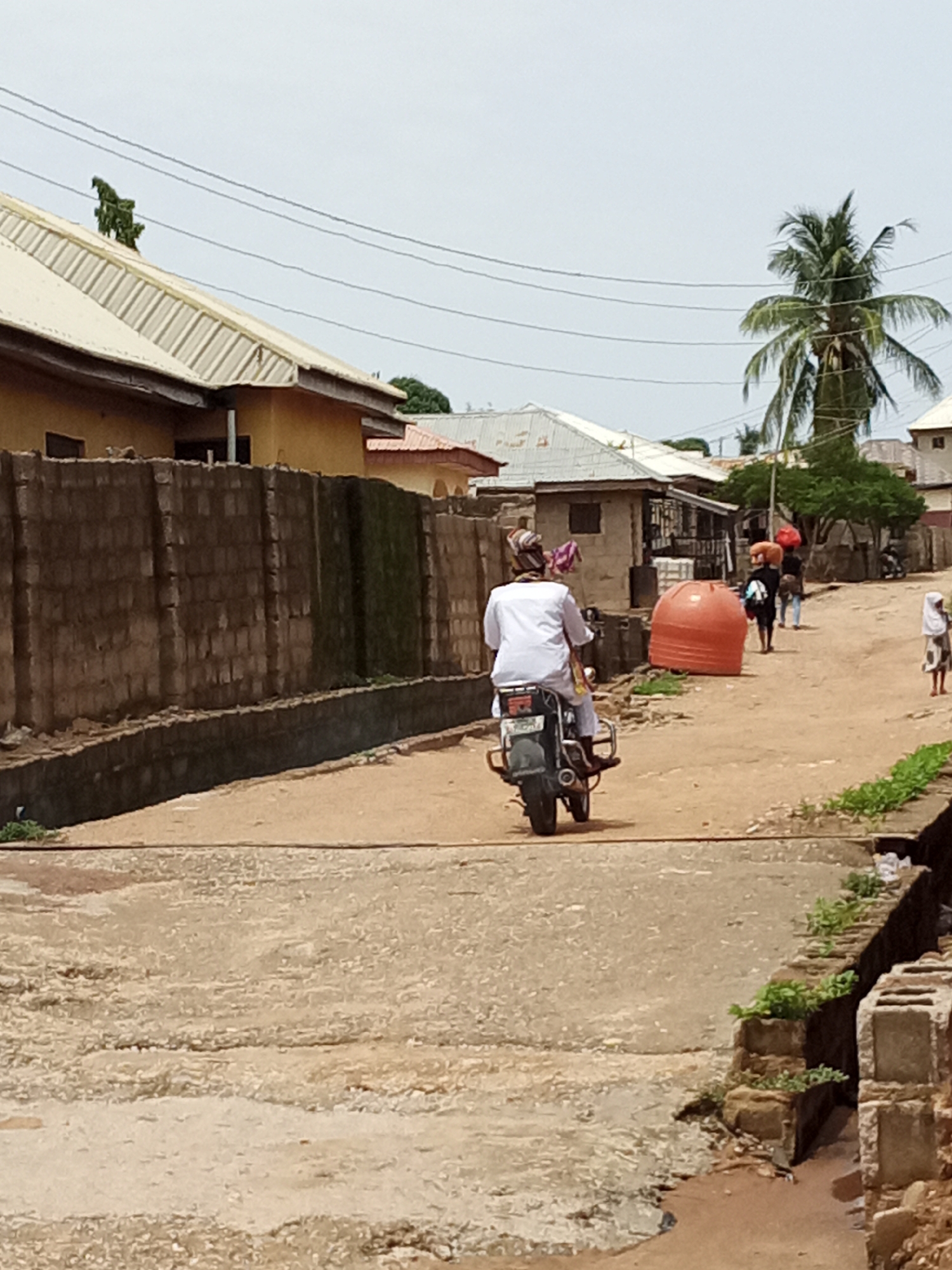 The image depicts a street scene in an urban area of Unknown, Unknown. In the foreground is a rough, uneven concrete road with a narrow channel or ditch running along the right side, lined with concrete blocks. On the road, further down the street, is a person driving a motorcycle away from the viewer. The rider is wearing a white shirt and what appears to be a head covering. Several other people are walking along the road in the same direction, carrying items on their heads. There is a large orange object, potentially a water tank, alongside the road. Buildings line both sides of the street, some with corrugated metal roofs and brick or concrete block walls. A palm tree rises above the buildings on the right side of the street. Power lines stretch across the sky above the scene.