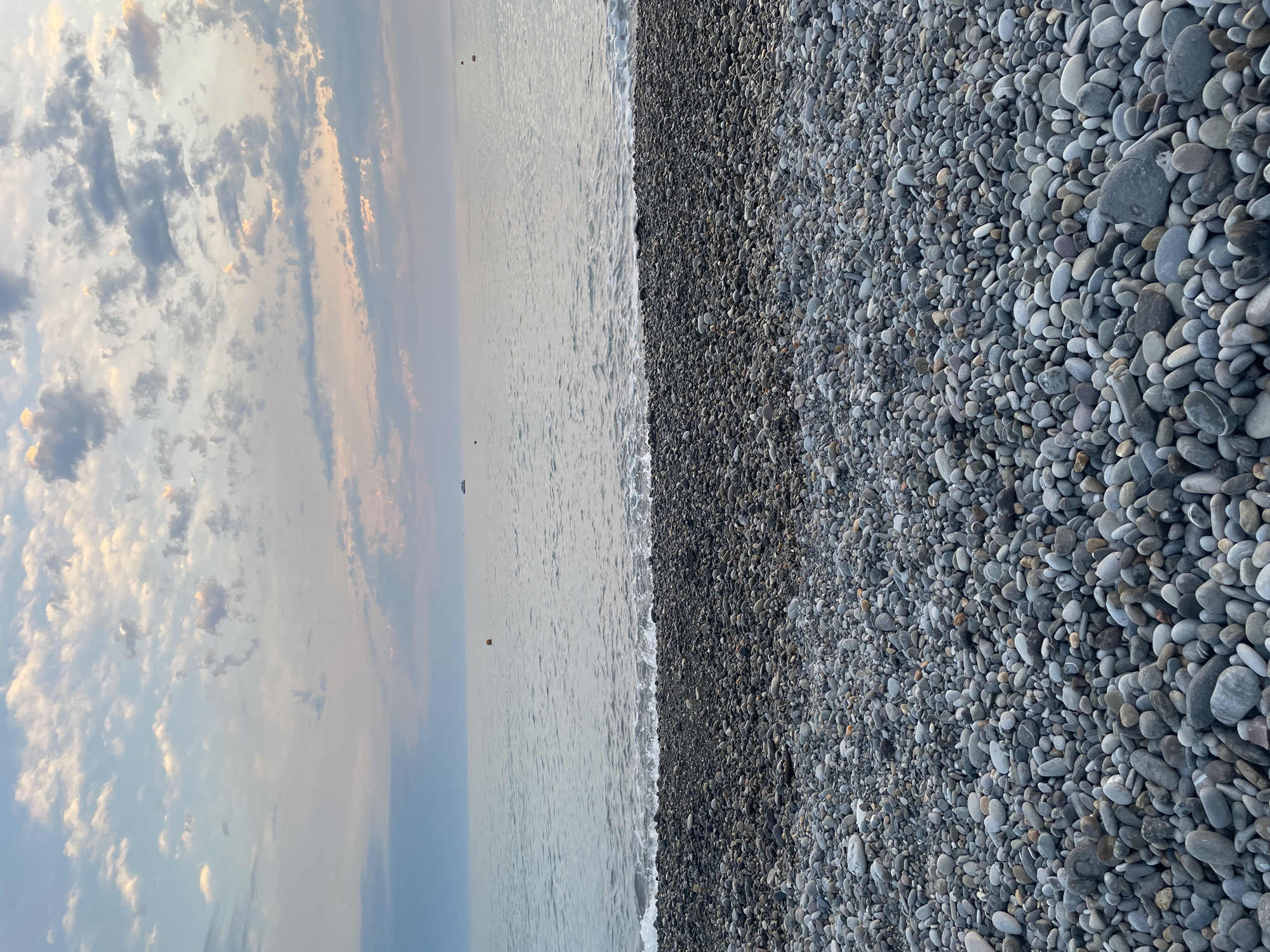 The image shows a rocky beach and the sea under a cloudy sky. The foreground is filled with smooth, grey, and dark-colored pebbles of various sizes, densely covering the ground. The sea extends to the horizon with gentle waves lapping at the shore. In the distance, there are a few buoys and a ship or boat. The sky is filled with soft clouds, with hints of pink and orange from the setting or rising sun, creating a serene atmosphere. The location is the city of Unknown, Unknown.