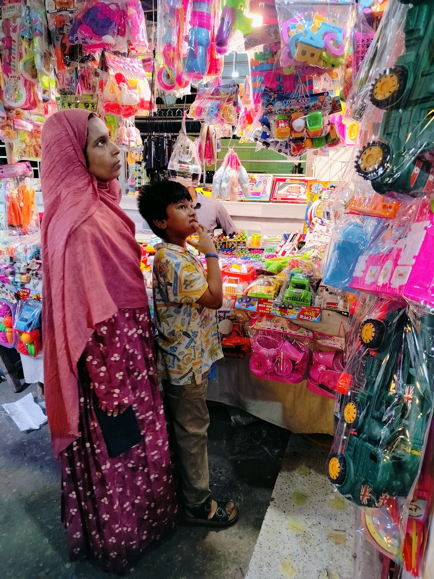 The image shows a woman in a pink hijab and a patterned dress standing next to a young boy. They are in a marketplace, likely in Dhaka, Bangladesh, with stalls displaying a variety of colorful toys and merchandise. The boy is looking upwards, seemingly interested in the toys hanging above. The woman is looking to her left, her expression unreadable. The stalls are packed with plastic toys, including toy cars, dolls, and building blocks, all packaged in plastic. The overall impression is a bustling, vibrant market scene.