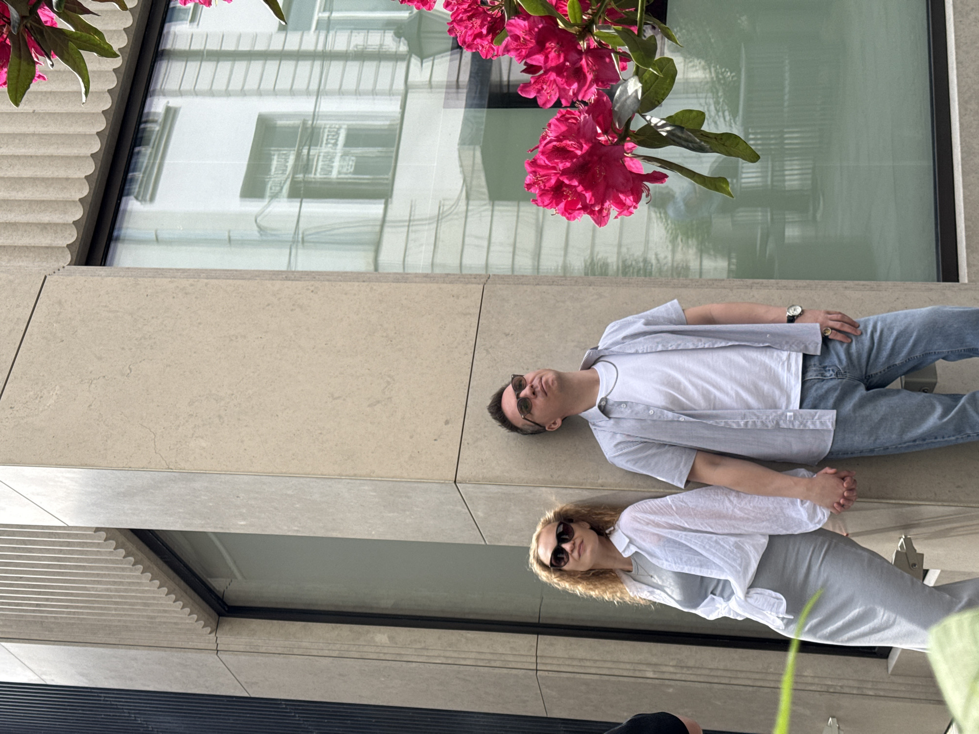 A man and a woman lie side by side on a beige stone surface, possibly a ledge or low wall, adjacent to a building with a mirrored window reflecting surrounding architecture. The man wears sunglasses, a white t-shirt under a light blue striped button-down shirt, and blue jeans. The woman also wears sunglasses and a white top with a light gray skirt or dress. Pink flowers are positioned above them, potentially hanging from a planter. The background shows a reflection of a building with rows of windows. Due to the architectural style and the presence of pink flowers, the location might be Moscow, Russia.