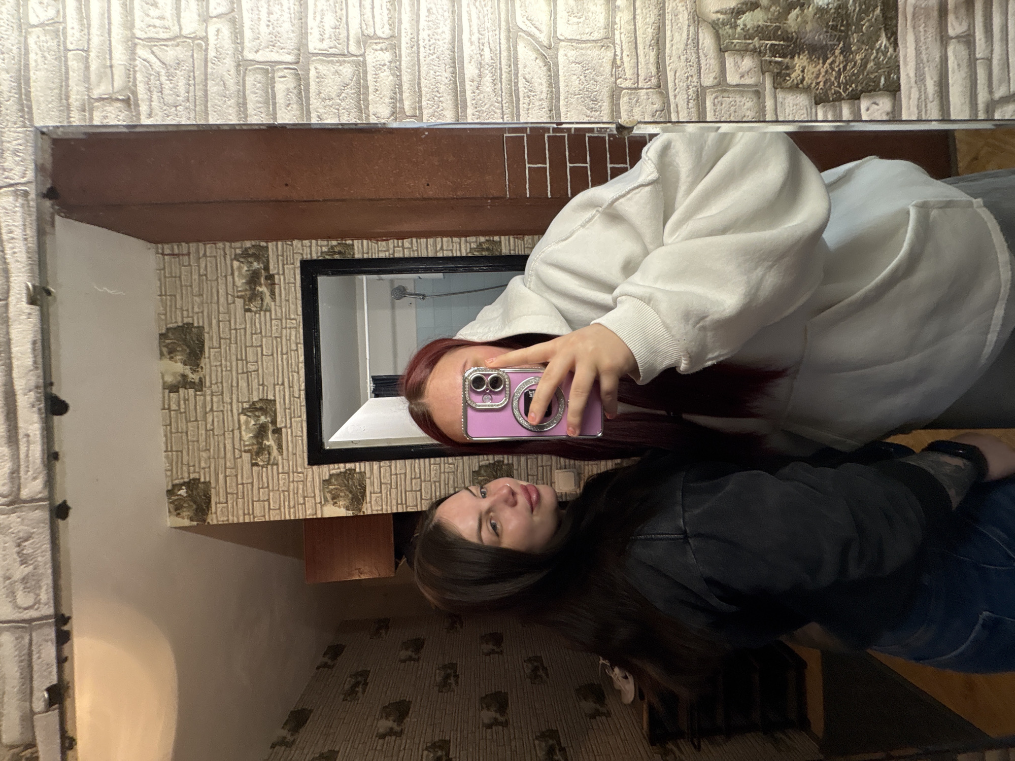 A selfie taken in a bathroom mirror. A person with auburn hair is taking a photo with a purple smartphone. The reflection in the mirror shows a second person with brown hair. The background features textured walls and a glimpse of a shower area. The location is assumed to be in Chornomorskoe, Ukraine.