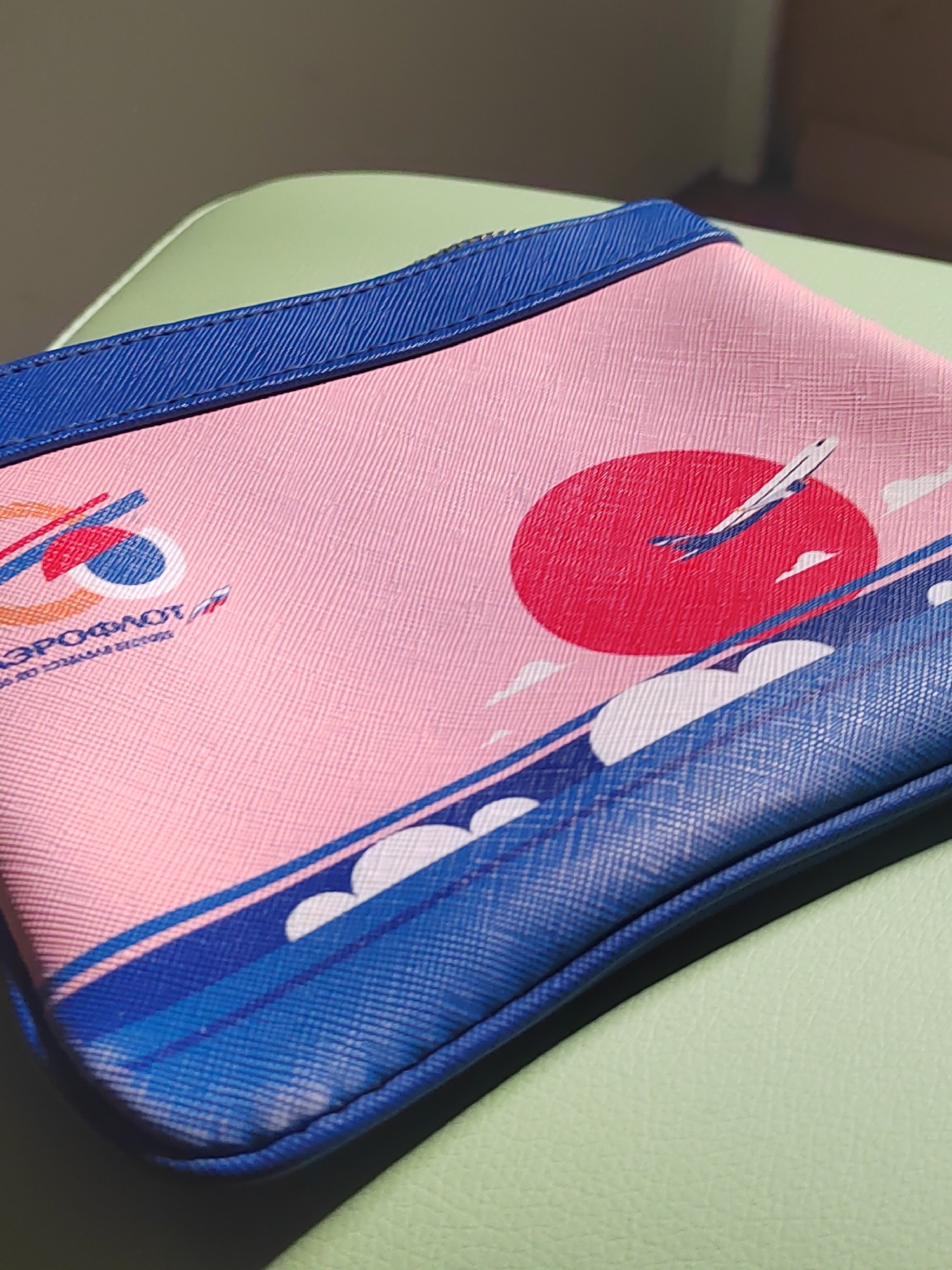 A close-up shot shows a pink cosmetic bag with a blue trim, featuring an Aeroflot airline design. The design includes the Aeroflot logo, an airplane silhouette against a red circle, and blue stripes with white cloud-like shapes. The bag sits on a light green surface. The city of Votkinsk, Russia, is not discernible in the image.