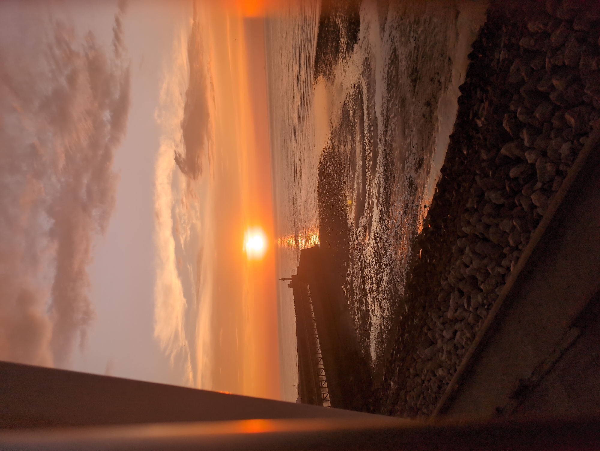 This image depicts a sunset over the sea at Le Tréport, France. The sun, a bright orange orb, is positioned low on the horizon, casting a warm glow across the sky and water. The sky is filled with soft, wispy clouds in shades of orange, pink, and purple. In the mid-ground, a long pier extends into the sea, leading to a lighthouse at its end. The pier is lined with what appear to be railings or fencing. The sea is relatively calm, with gentle ripples reflecting the colors of the sunset. In the foreground, the shore is covered with rocks and wet sand, suggesting that the tide is out. A portion of a railing or barrier is visible on the left side of the frame, obscuring part of the view. No people are clearly visible in the scene, though there may be small figures on the pier. The overall atmosphere is serene and picturesque, characteristic of a coastal sunset.