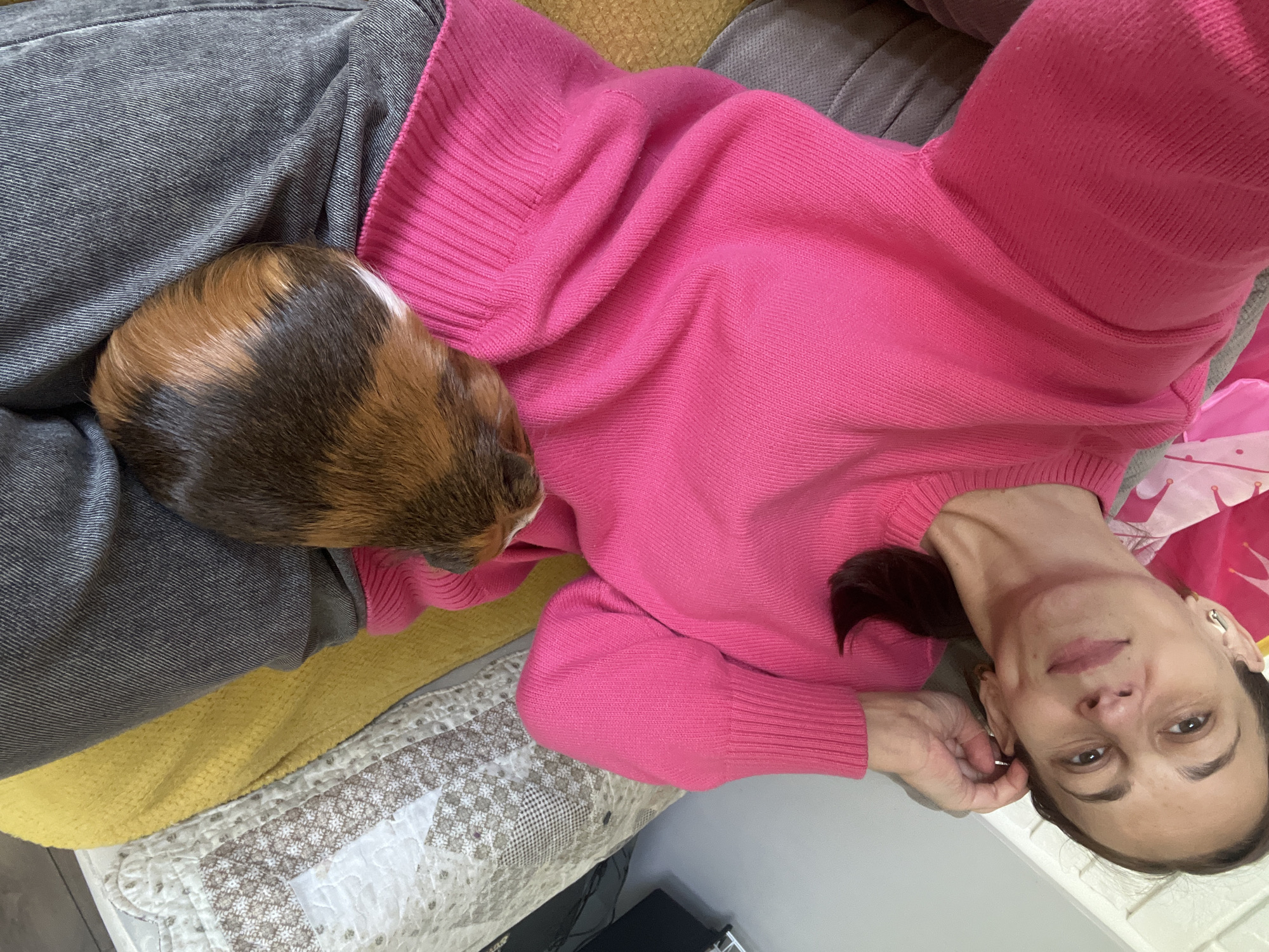 An overhead, medium shot shows a woman lying on a couch, wearing a bright pink sweater and gray pants. She is looking up at the camera with a slight smile. Her left hand is resting on her head, and her right arm is draped across her body. A guinea pig with brown, black, and white fur is lying on her abdomen, its head facing to the right. The background shows a couch with a yellow blanket and a white and brown patterned quilt.