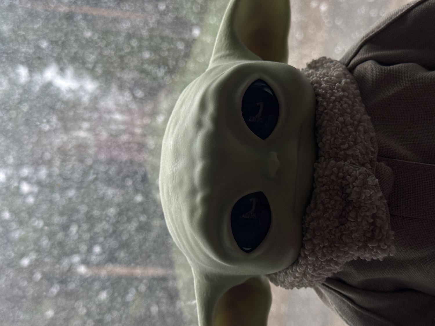 The media file presents a close-up portrait of a toy resembling Grogu (Baby Yoda) from The Mandalorian, oriented on its side with its head facing left. The subject's light green face features large, dark, and reflective eyes, giving it a calm or observant expression. Subtle textures and gentle contours are visible on its skin. Large ears extend from the sides of its head.
The toy is dressed in a light brown, sherpa-like textured collar that appears soft and fluffy, transitioning into a simpler, light brown, woven fabric outfit covering its visible body.
The background is heavily blurred, characterized by numerous small, circular bokeh highlights. This effect suggests diffused light reflecting off a surface, possibly water droplets on a windowpane, creating a soft, dreamlike backdrop in shades of grey, green, and white. The lighting is soft and ambient, illuminating the toy from the front and slightly to the right, creating gentle shadows that add depth to its features and clothing.
The scene conveys a quiet and contemplative mood. No specific activity is depicted, and there is no visible text. The setting appears to be indoors, likely positioned near a window during an overcast or rainy day, as indicated by the bokeh in the background.