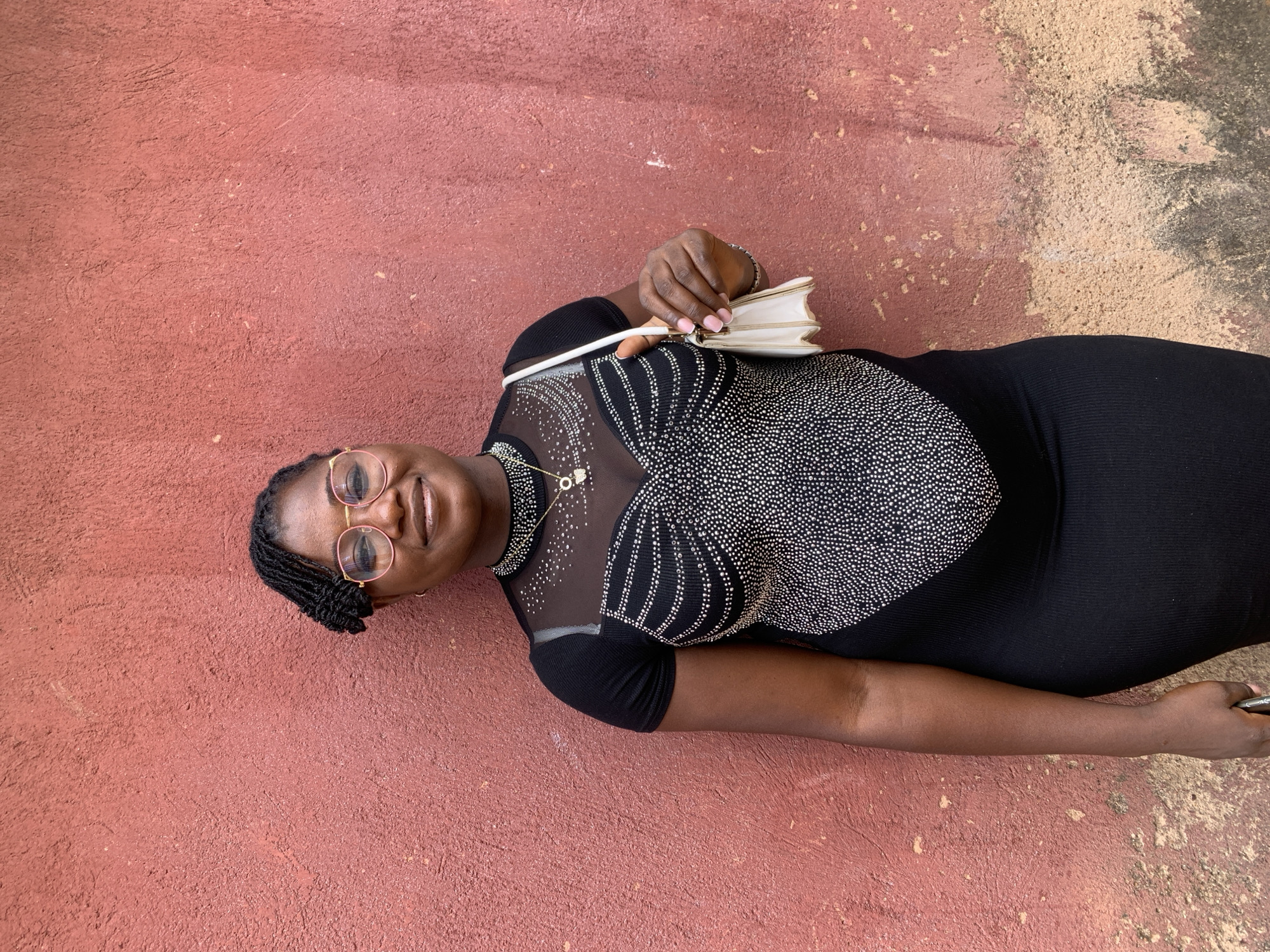 A woman wearing glasses and a black dress poses on a surface that transitions from red to a lighter, textured color. She holds a small white clutch. The location is not specified and cannot be determined from the image.