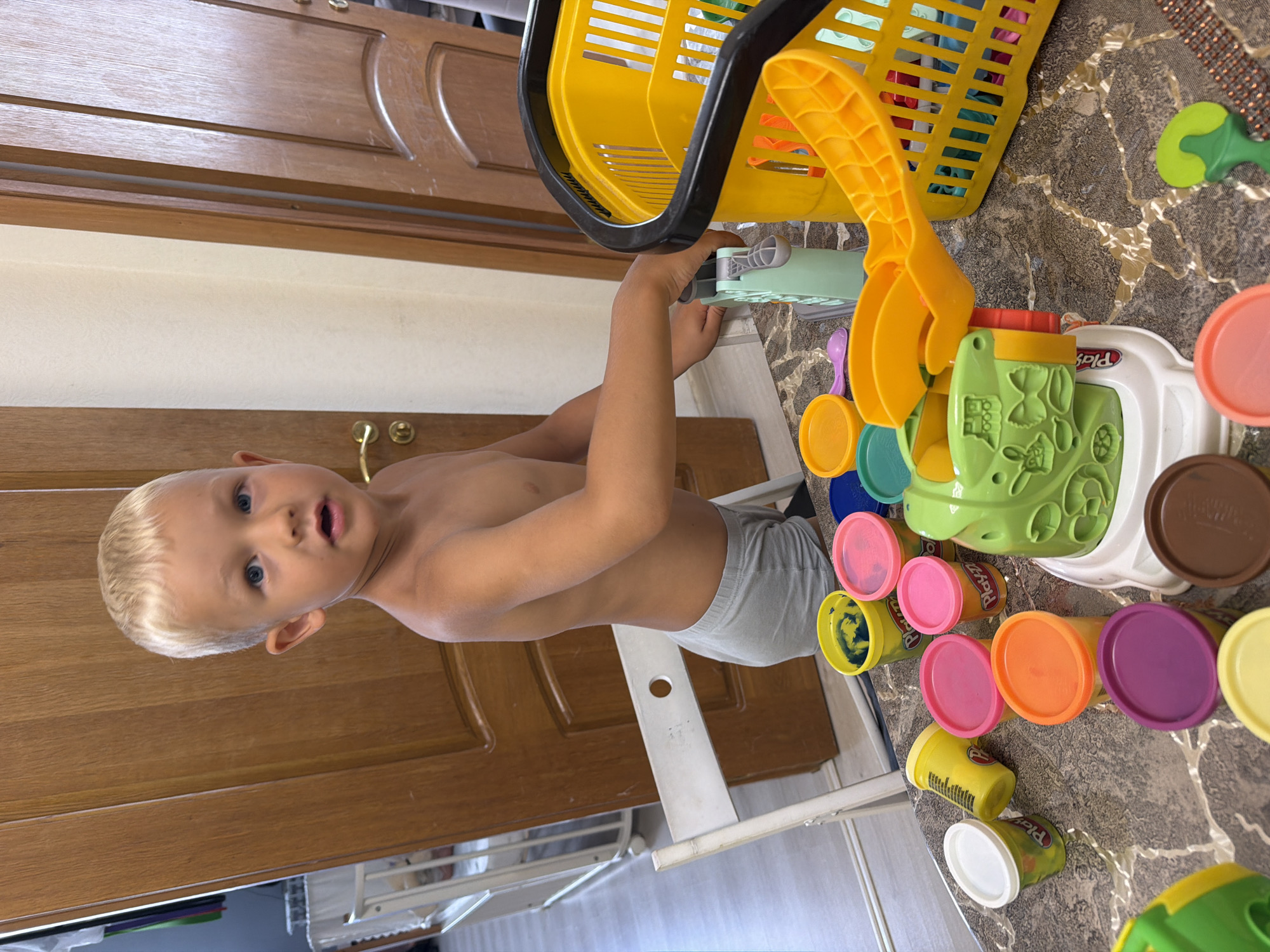 A young, fair-haired boy, shirtless and wearing grey shorts, stands in a room with wooden doors. He is positioned behind a table covered with various colorful Play-Doh containers and a Play-Doh set with green and white components, along with a yellow basket. The boy is looking towards the camera with an open mouth. The room appears to be a domestic setting, possibly a playroom or bedroom, with a crib visible in the background. The flooring is light-colored with a subtle pattern, and the table has a marble-like finish. The scene is brightly lit, suggesting it is daytime. There is no information in the image to indicate the city of Sochi, Russia, or any other specific location.
