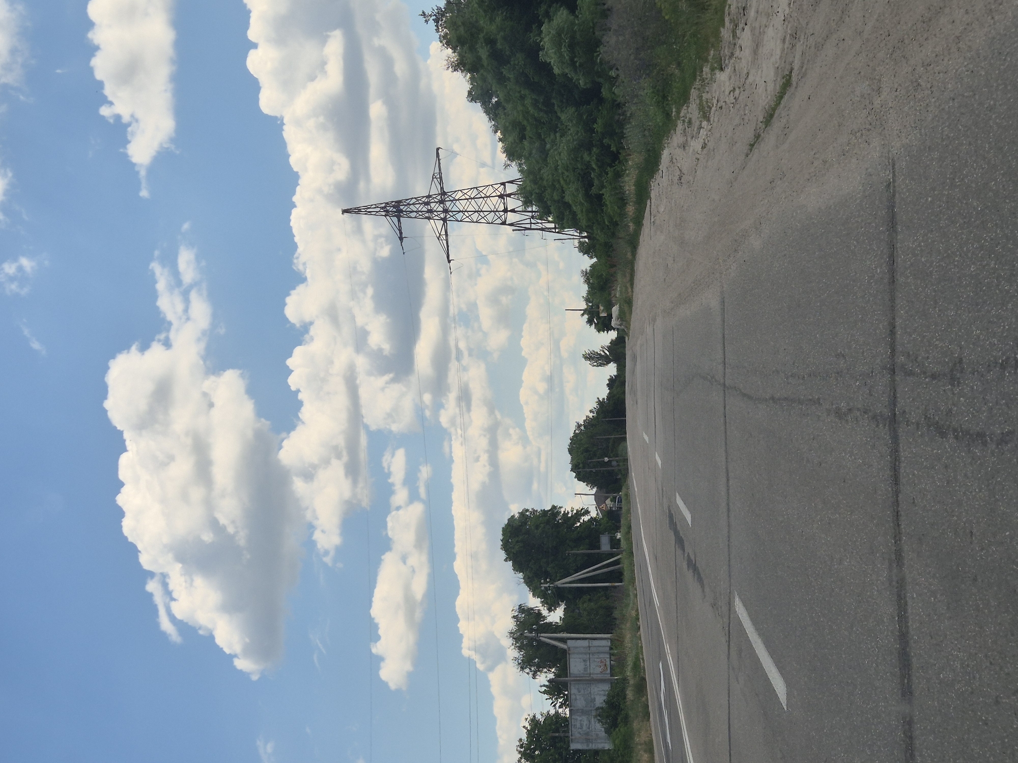 An eye-level, medium-distance shot captures a paved road stretching towards a horizon dominated by a clear blue sky with scattered white clouds. The road, marked with white dashed lines, curves slightly to the right, bordered by a dusty shoulder on the right and grassy verges on the left. In the background, a tall, latticed electrical pylon stands prominently against the sky, surrounded by dense green trees and vegetation. To the left of the road, several concrete poles support overhead wires, and a large, weathered billboard is visible. The scene suggests a rural or semi-urban setting in Ukraine, potentially in or near the city of Pidhaitsi. There are no people or vehicles visible in the frame.