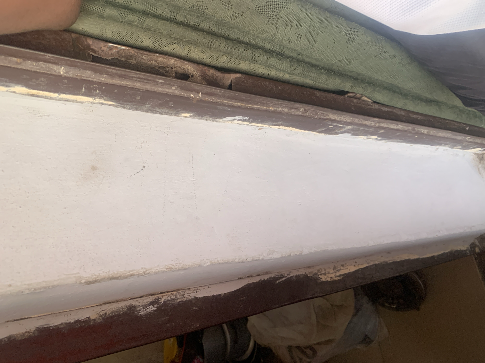 This is a close-up, eye-level, vertical, medium shot of a weathered wooden door frame and the adjacent plastered wall. The door frame is dark brown with chipped paint, revealing lighter wood underneath. A thin strip of what appears to be a metal frame is visible on the right side of the door frame, partially obscured by a green patterned curtain. The plastered wall is painted white but shows signs of dirt, scuff marks, and peeling paint, particularly along the edge of the door frame. The background to the left shows a dark interior with some white fabric or plastic sheeting and possibly some metal pots or containers. To the right, the green curtain hangs down, and a white mesh fabric is visible at the bottom right corner. The lighting suggests an indoor environment, possibly a home or a less maintained building, in Jauro Yinu, Nigeria. The scene appears to be a corner of a doorway, emphasizing the condition of the infrastructure.