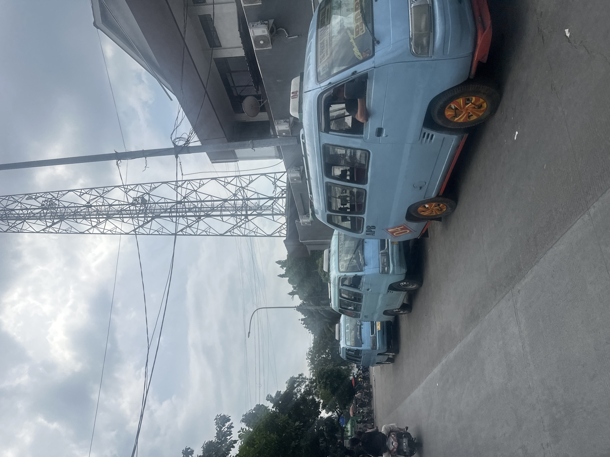 The image shows a street scene in North Jakarta, Indonesia. Three light blue public transportation vans, likely "Angkot" or "Mikrolet" vehicles, are parked in a row along the side of the road. The first van has a partial view of the driver. The wheels of the vans are orange and red. A large metal utility pole stands on the left side of the image. The sky is overcast. A building is above the vans. Trees are visible in the background. A motorbike and other vehicles parked behind the vans. The vans each bear a number.