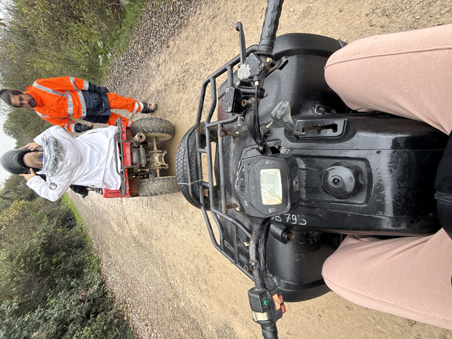 An overhead, first-person view depicts a line of all-terrain vehicles (ATVs) on a dirt path in Çiftlikköy, Turkey. In the foreground, the lower torso and legs, clad in light brown sweatpants, are visible from a seated position on a black ATV. The handlebars, control panel with a display, and a front cargo rack with a secured spare tire are visible on this ATV. Directly ahead, a red ATV is positioned, facing away. A person wearing a white winter jacket with a fur-lined hood is seated on the red ATV, hands raised near their head, seemingly adjusting a dark hood or helmet. The red ATV bears the alphanumeric identifier "LABR 041" on a rear plate. To the right of the red ATV, a male individual stands on the dirt path. He wears a bright orange high-visibility jacket and trousers with dark blue accents, and work boots. He has dark hair tied in a bun and a beard, and is looking towards the person on the red ATV. The path is unpaved, appearing damp and muddy, bordered on both sides by dense green foliage and trees under an overcast sky.