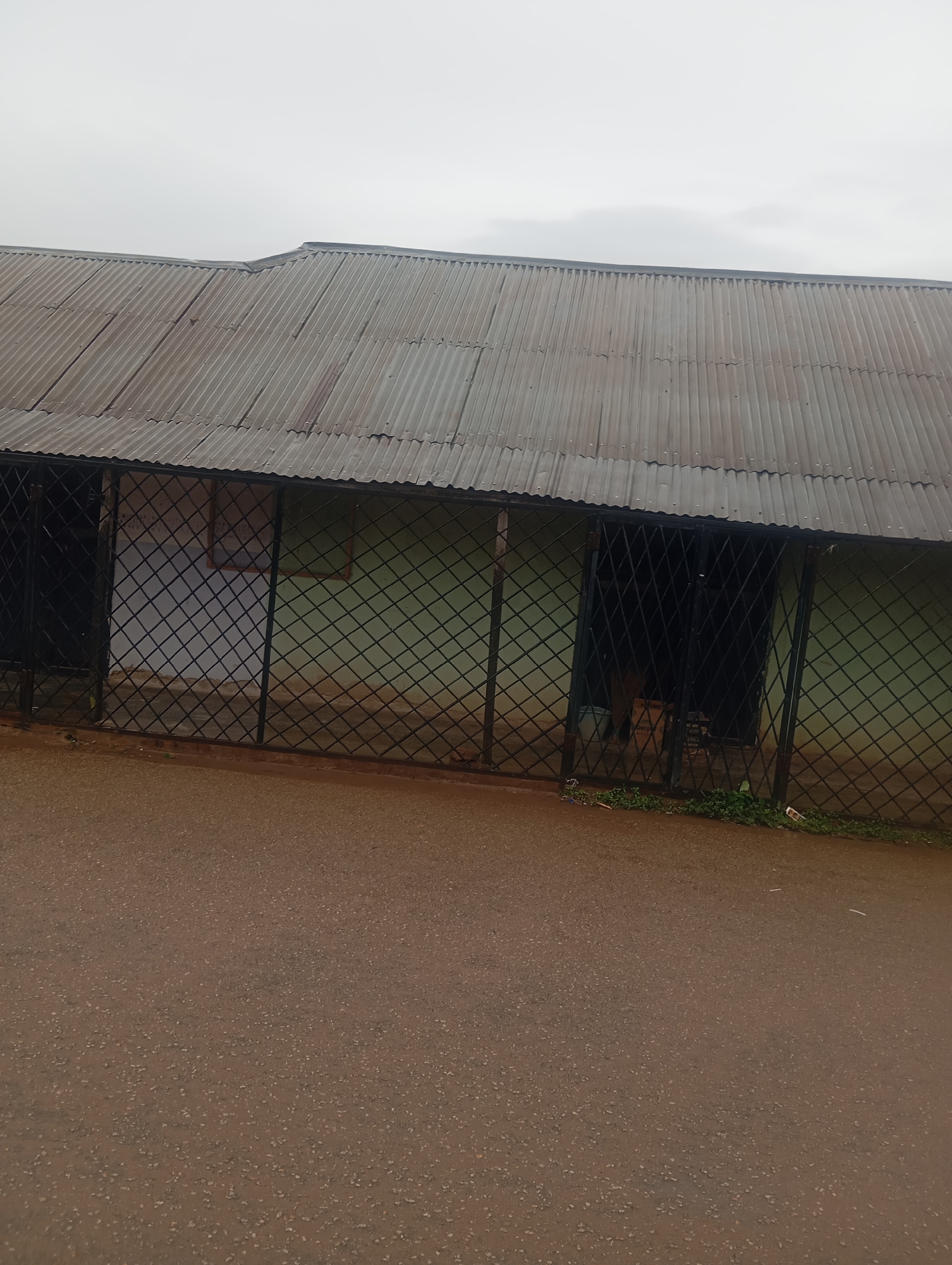 The image shows a light green building with a corrugated metal roof, likely in Jos, Nigeria. A black metal grate fence runs along the front of the building, obscuring a portion of the entrance and windows. The building appears to be single-story, with a slightly uneven roofline. The foreground is a paved surface, possibly a road or pathway, with scattered small stones. The sky is overcast, suggesting a cloudy day. There are no people visible in the image, and the overall scene appears to be an exterior view of a utilitarian structure.