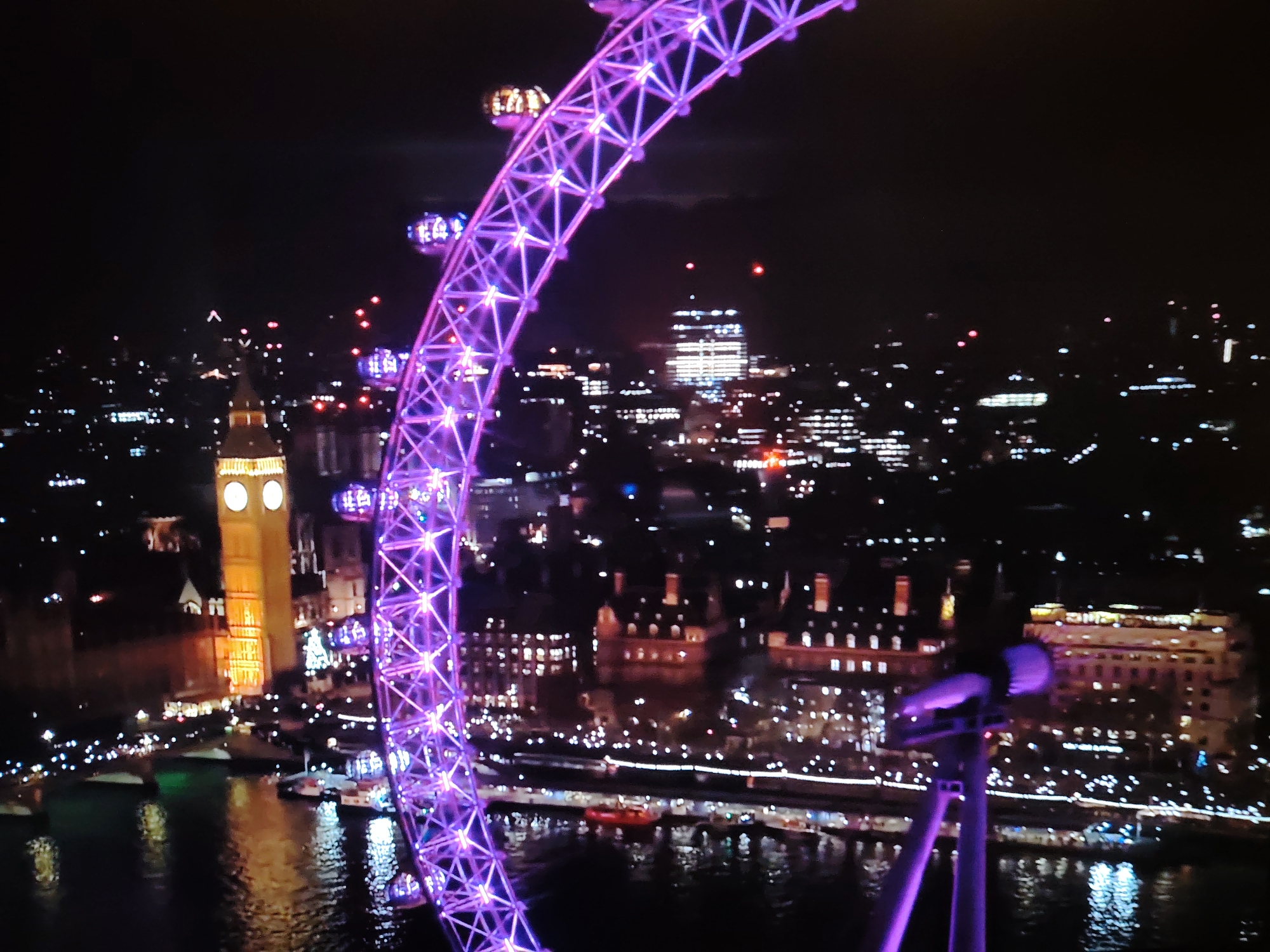An aerial nighttime view captures the illuminated structures of London. In the foreground, a portion of a brightly lit purple Ferris wheel, the London Eye, dominates the right side of the frame. The iconic Elizabeth Tower, commonly known as Big Ben, stands to the left, its clock face glowing orange. The Thames River flows in the middle ground, reflecting the city lights. Numerous buildings with illuminated windows form the urban landscape stretching into the background. The scene is set against a dark night sky, suggesting a clear evening.