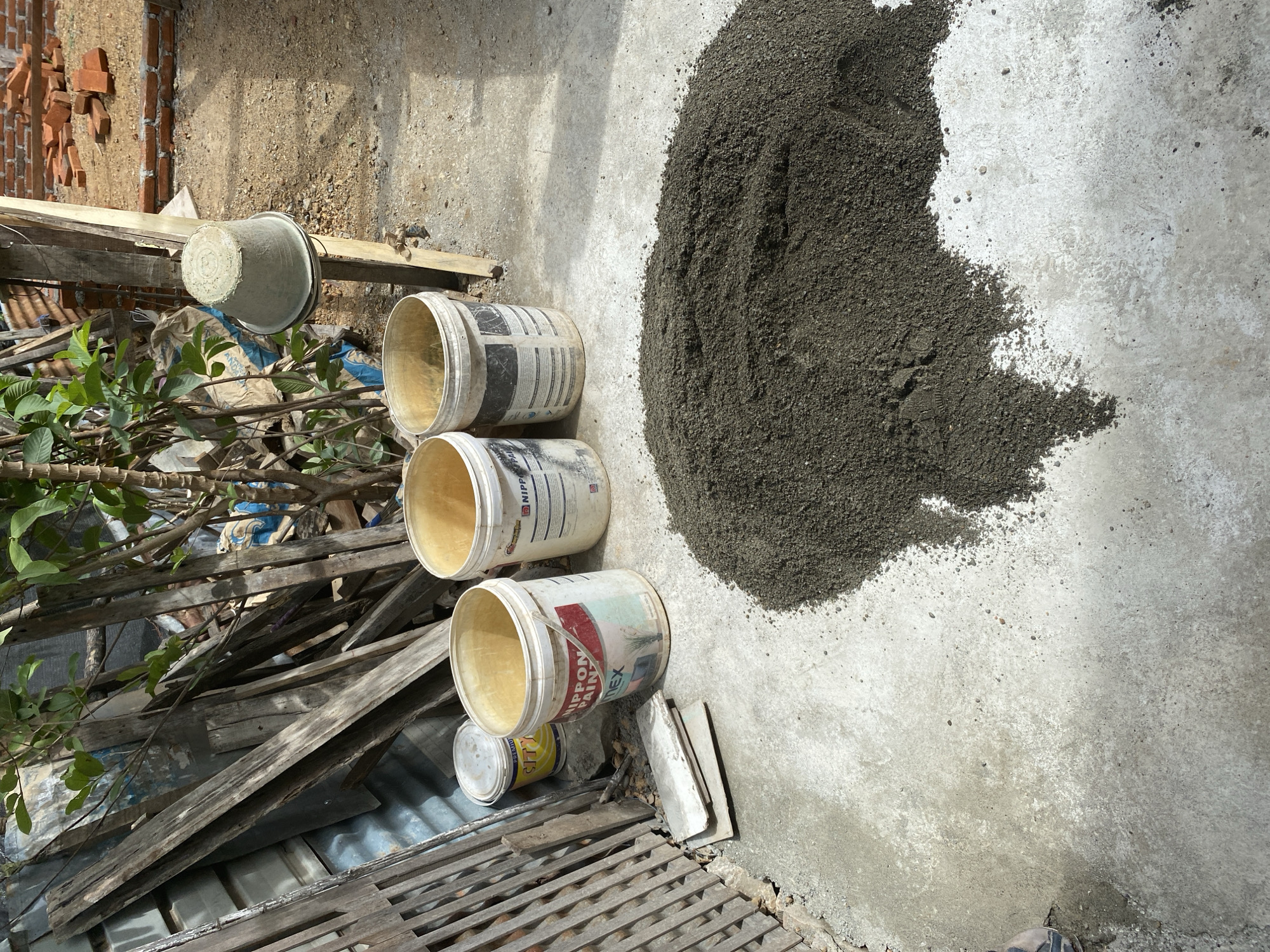 An exterior shot shows a construction site with a pile of dark, granular material, possibly cement or sand, on a light gray concrete surface. Three white plastic buckets are arranged in a line on the concrete to the left of the pile. One bucket, on the far left, is smaller and has a yellow label. The other two buckets are larger and appear to be the same brand, with red and black text. Behind the buckets, a stack of weathered wooden planks leans against a structure. A light gray plastic bucket hangs from a wooden post in the background. Further back, a pile of red bricks is visible on a dirt surface. The scene is illuminated by natural sunlight, casting shadows on the ground. The overall impression is one of a construction or renovation project in progress.