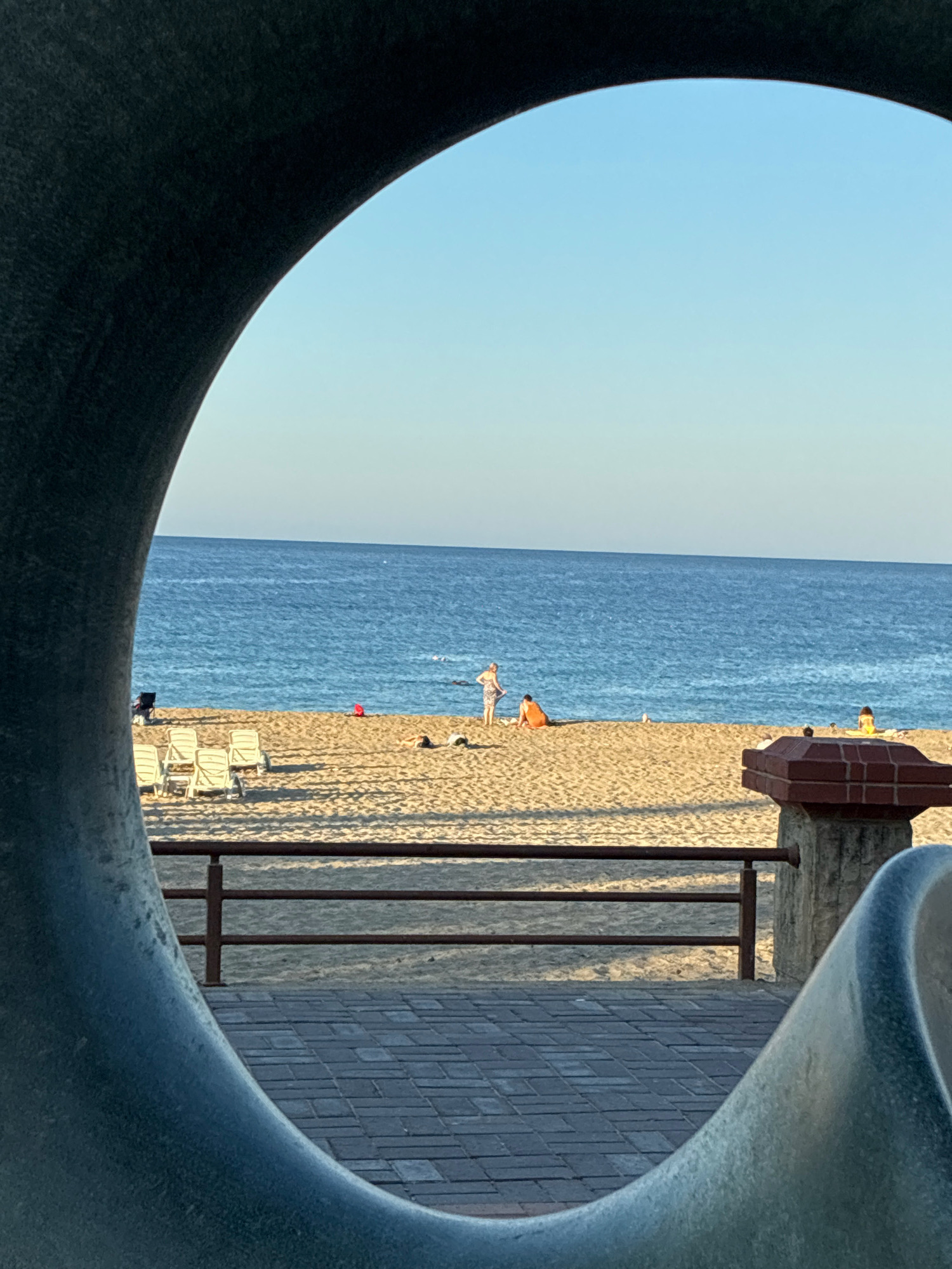 This is a medium shot, eye-level, outdoor photograph taken at a beach in Alanya, Turkey. The scene is framed by a large, dark, circular metal sculpture in the foreground, which partially obscures the view of the beach and the sea beyond.
The beach is sandy and appears to be a public area, with several empty lounge chairs lined up in the sand on the left side of the frame. Further back on the beach, there are a few people. One person, wearing a light-colored top and patterned shorts, stands near the water. To their right, two people are seated on the sand; one is wearing an orange shirt and the other is lying down. A red beach umbrella is visible in the sand behind the seated individuals. There are also a few other people scattered further down the beach.
Beyond the sandy beach, the blue sea stretches to the horizon, meeting a clear, light blue sky. The water appears calm with gentle waves. A dark-colored railing runs across the middle of the frame, separating the paved walkway from the beach. The walkway is paved with grey rectangular bricks. On the right side of the frame, a section of a stone pillar with a dark red brick cap is visible. The overall lighting suggests it is late afternoon or early morning, with soft sunlight casting shadows on the sand.