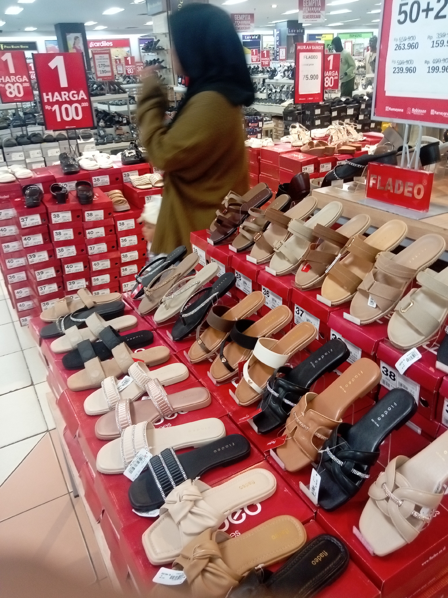 The image shows a display of women's sandals for sale at a department store in Batam, Indonesia. The sandals are arranged on red display boxes, with various styles and colors visible. A woman wearing a headscarf and a brown jacket is browsing the sandals in the foreground. In the background, other shoppers and displays of shoes can be seen. Signs indicate prices and brand names like "Fladeo." The overall environment is a retail store with a tiled floor and brightly lit displays.