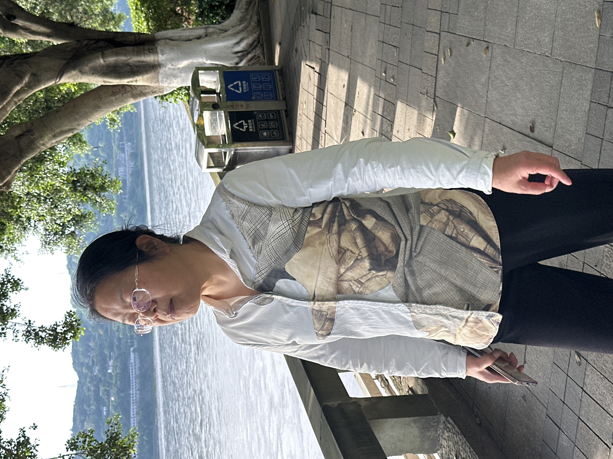 The photograph shows a woman with short, dark hair and glasses, standing near a body of water with trees and greenery visible on the opposite shore. She is wearing a light-colored, patterned shirt and dark pants, and is holding a mobile phone in her right hand. A tree and recycling bins are located behind her on the paved path. The scene appears to be a park or waterfront area in Jiangjin, China.