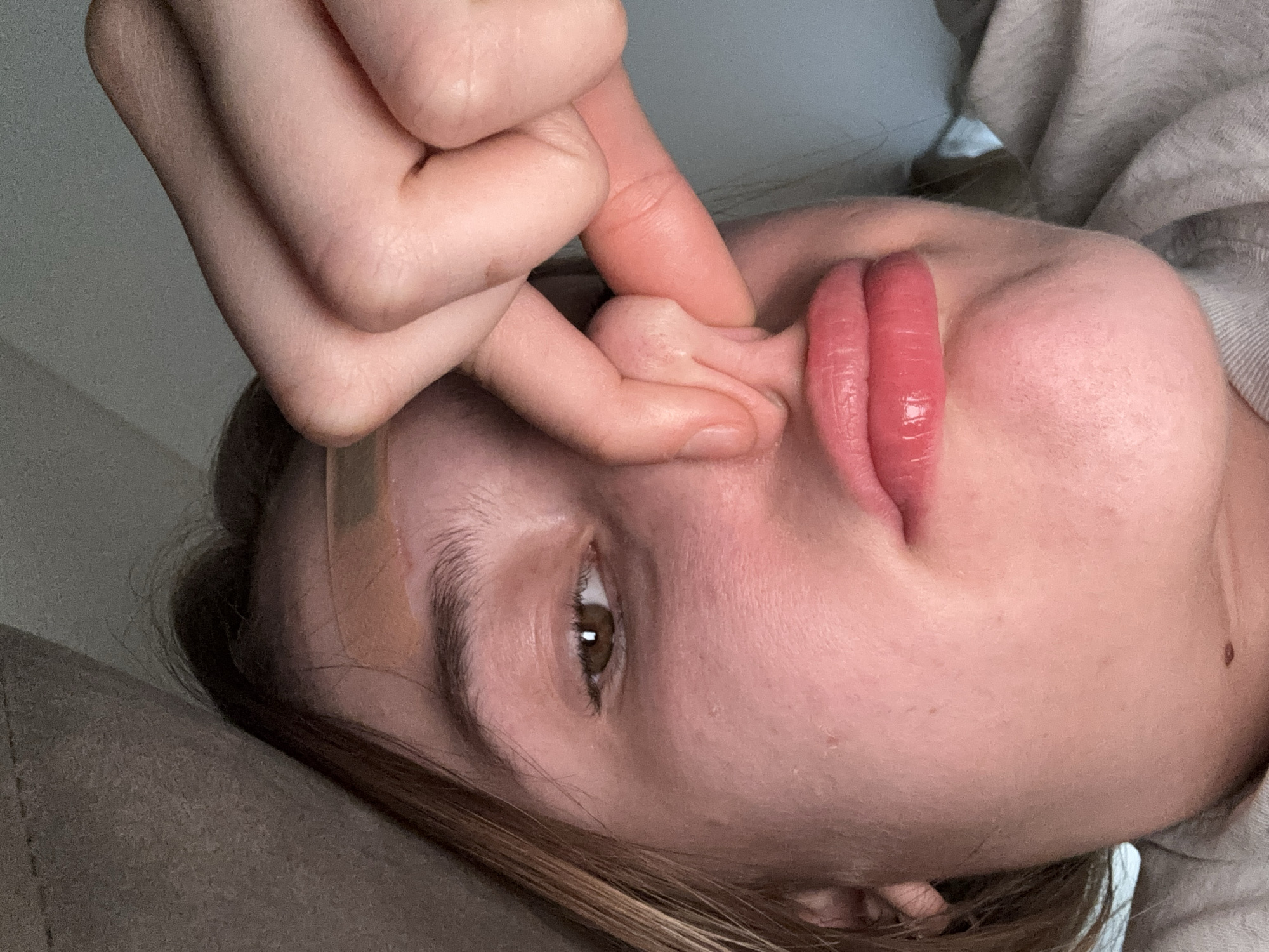 A close-up shot shows a young woman's face. Her left eye is visible, with light brown irises and dark lashes. She has plump, pink lips and her nose is being pinched by fingers from the right side of the frame. A light-colored band-aid is visible on her forehead, just above her eyebrow. The background is blurry and appears to be indoors, with fabric elements suggesting a couch or bedding. The lighting is dim, with some bright spots on her skin. There is no information in the image that suggests a location in Lednevo, Russia.