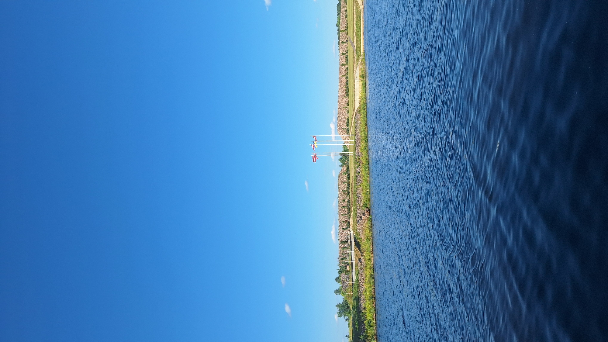 A bright, sunny day with a clear blue sky over a calm, dark blue body of water. In the distance, a low, stone wall forms a raised embankment. A bridge crosses a gap in the embankment, and beyond it, four flags on tall white poles stand against the wall. The flags appear to be national flags, one of which is red with a white stripe. Trees and green grass line the embankment, and in the far distance, more trees are visible. The overall scene suggests an outdoor recreational area or park setting. The location is identified as unknown, Latvia.