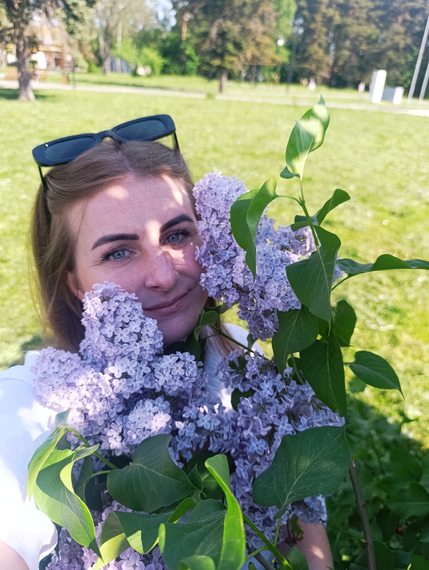 A medium outdoor shot shows a fair-skinned woman holding a bouquet of purple lilacs. The woman faces the camera with a gentle smile, her blue eyes and freckled complexion visible. Dark sunglasses are perched atop her light brown hair, which is styled in a ponytail. The bouquet of lilacs is substantial, with several large clusters of flowers obscuring part of her face and body. Green leaves provide a backdrop to the blooms. The background features a grassy park with trees visible in the distance. The scene is bathed in bright, natural light.
