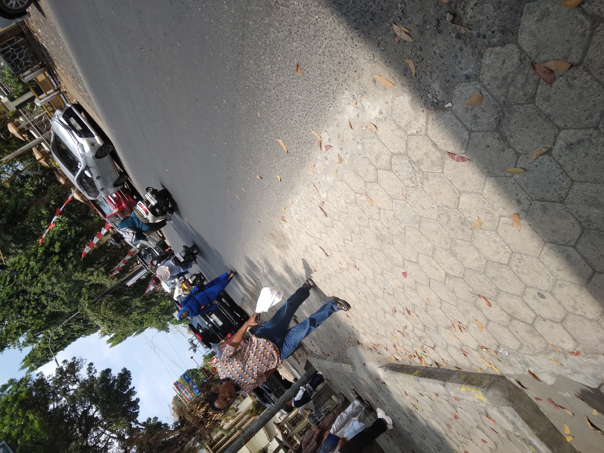 The image depicts a street scene in Desa Pamijen, Kecamatan Sokaraja, Indonesia, on a sunny day. A man in a patterned short-sleeved shirt and blue jeans walks on the sidewalk carrying a white plastic bag. The sidewalk is paved with hexagonal tiles and has fallen leaves scattered on it. To the man's right, a paved road is visible with several vehicles, including a silver minivan, a red car, and a black SUV. A woman in a blue headscarf and outfit is walking on the road near the black SUV. Another woman on a scooter is also on the road. The background features trees, buildings, and a blue sky with scattered clouds. Red and white flags are visible along the roadside.