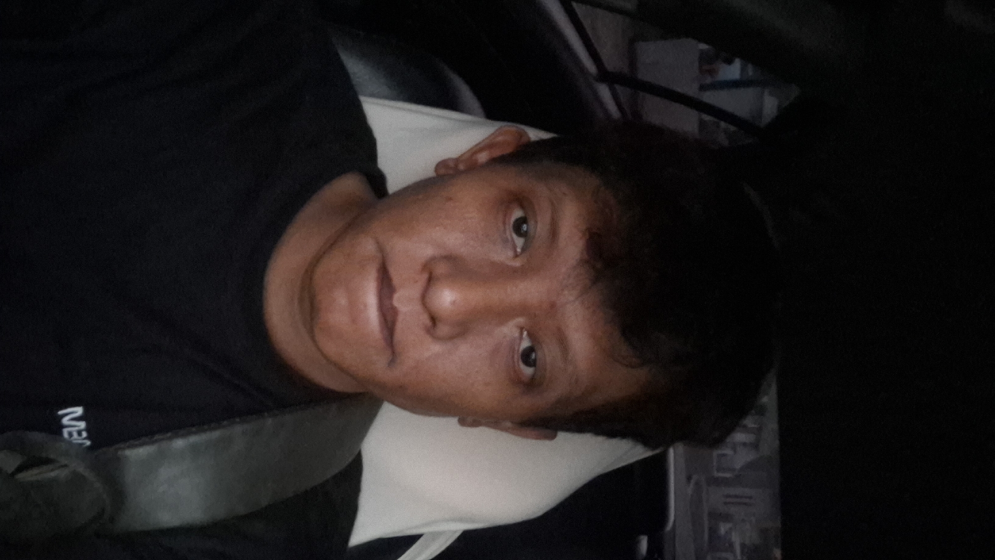The image is a close-up, selfie-style photograph of an adult male with dark hair and medium-toned skin. He is wearing a black t-shirt and a seatbelt is visible across his chest. The subject is inside a vehicle, indicated by the presence of a headrest and the interior of a car. The background is out of focus and suggests an outdoor setting with buildings visible, possibly a residential area. The lighting is dim. The location is Ciharashas, Indonesia.