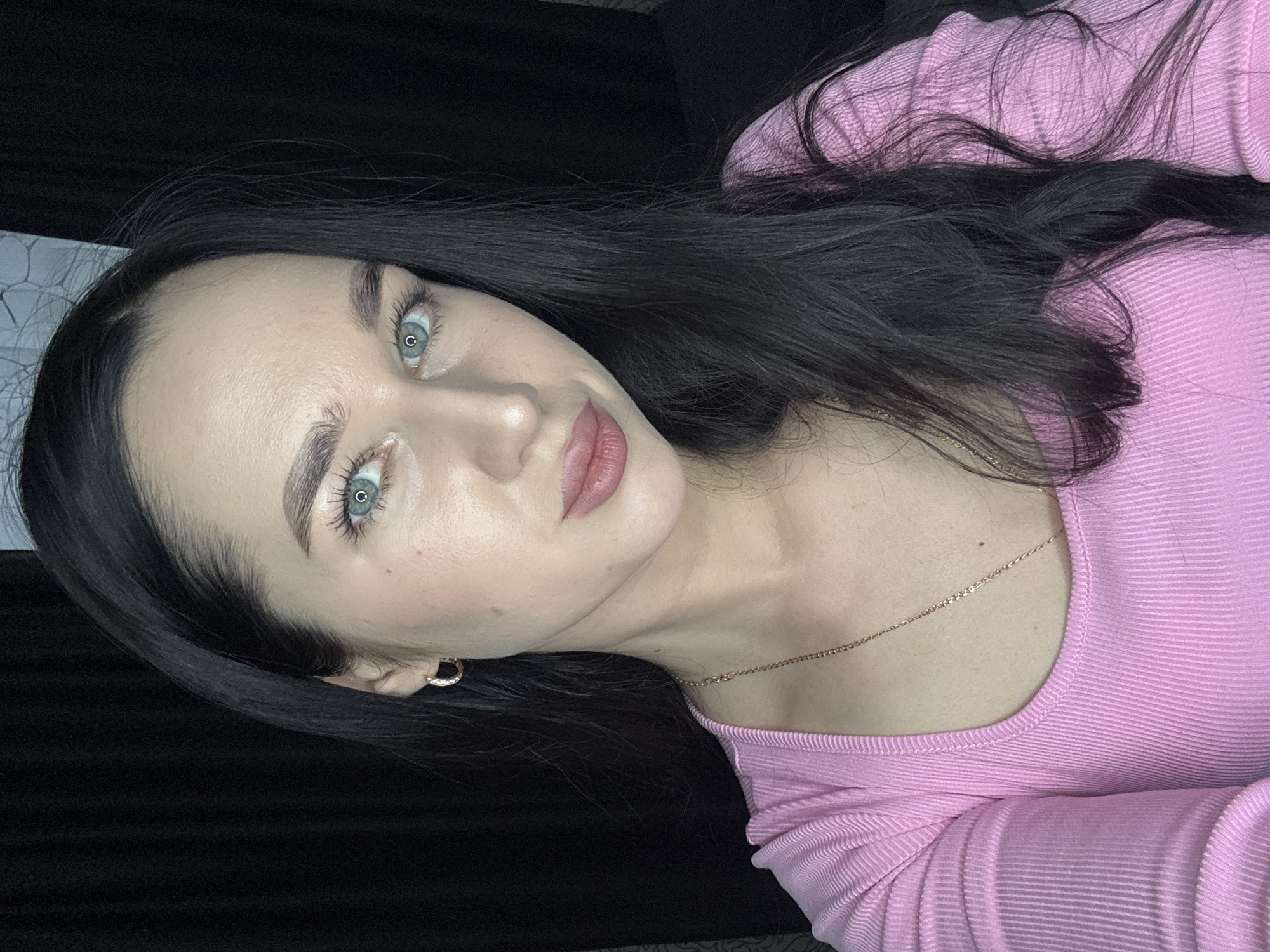 The image is a close-up portrait of a woman with long, dark hair and light blue eyes. She is wearing a pink, ribbed, long-sleeved shirt and a delicate gold chain necklace. She has on a small hoop earring in her left ear. The background is dark and appears to be a curtain or fabric. There are no discernible elements in the image that can identify the location as Novosibirsk, Russia.