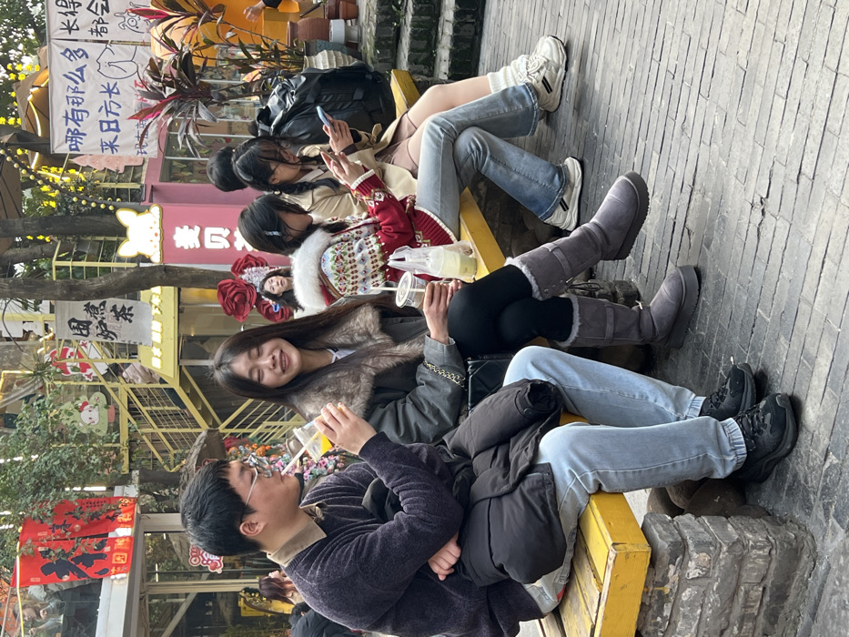 The scene is an outdoor pedestrian area within the city of Liangjiang, China. Four individuals are seated left-to-right on a yellow wooden bench supported by stone blocks. On the far left, a male wearing glasses, a dark purple sweater over a light brown collared shirt, and light blue jeans sips from a clear plastic cup containing an orange beverage through a straw. A black jacket is folded across his lap. Next to him, a female with long dark hair, dressed in a grey jacket with a fur collar, black leggings, and grey tall boots, holds a clear plastic cup with a light-colored beverage and smiles with her eyes partially closed. To her right, another female in a red and white patterned sweater with a fur-lined hood and light blue jeans is looking down at a smartphone in her hands; a clear plastic cup and a plastic bag are positioned beside her. On the far right, a fourth female, partially visible, wearing a light brown jacket and light-colored shorts, is also focused on a smartphone. A black backpack rests next to her.
The background consists of multi-level commercial structures featuring yellow metal staircases, numerous Chinese signs including red banners, and a large pink building with a white animal head silhouette. Decorative elements include red floral installations, hanging greenery, and various small cartoon cutouts. The ground is paved with rectangular grey stones.
