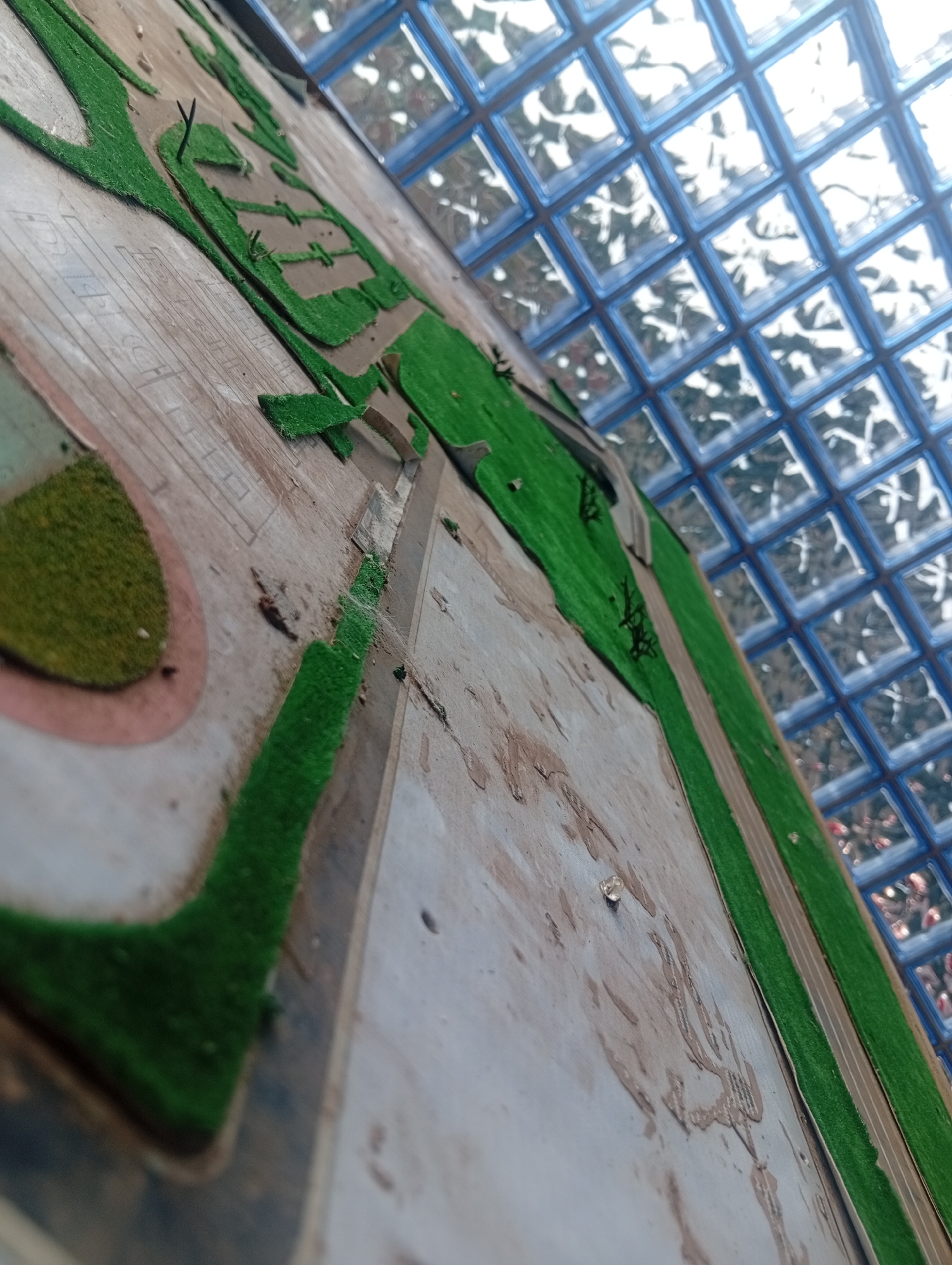 This is a close-up, high-angle shot of a diorama, likely depicting a section of Ukukwa, Nigeria. The diorama features artificial green turf representing grass, laid out in various shapes, including pathways and what appears to be a park or recreational area. The ground of the diorama is a light brown color, with some textured lines and splotchy markings that could represent roads or other paved surfaces. In the background, a grid of blue-tinted glass blocks is visible, likely part of the building's ceiling or a skylight. The lighting suggests an indoor environment with natural light filtering through the glass blocks. There are no people or animals present in the diorama.