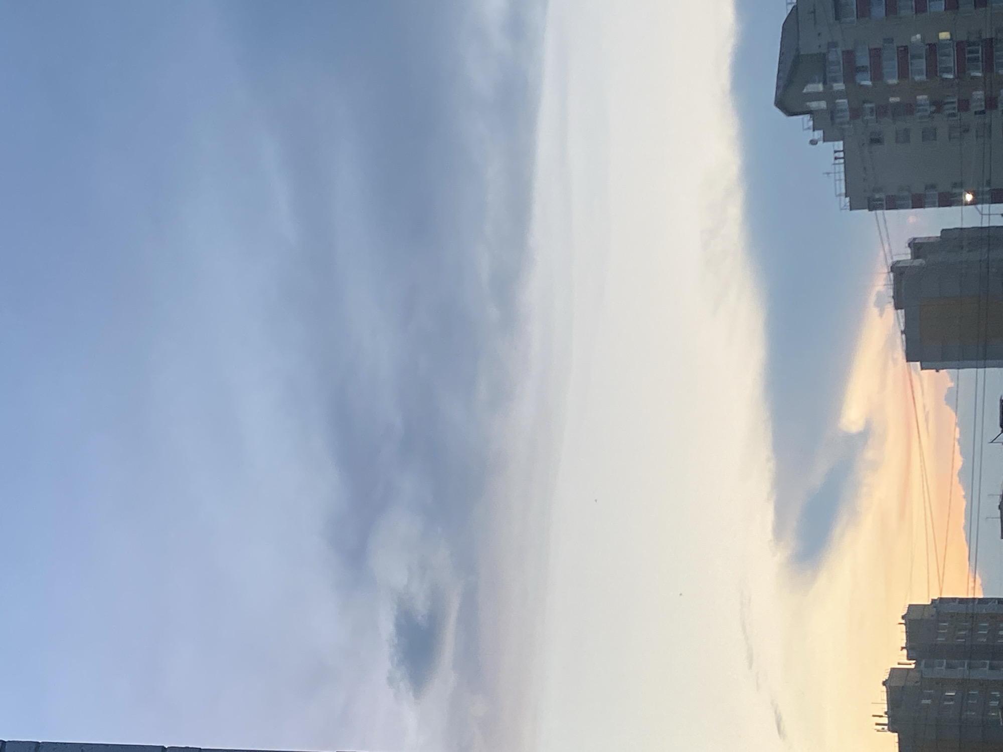 The image shows a view of apartment buildings against a hazy sky. The sky is a gradient of light blue and white with streaks of darker gray clouds scattered throughout. Towards the bottom of the frame, the sky transitions to a soft orange and pink hue, suggesting either a sunrise or sunset. Two tall apartment buildings are visible on the right side of the image, with several smaller buildings on the left. The buildings appear to be residential, with many windows visible. Electrical wires are strung between some of the buildings. The overall scene is overcast and somewhat muted in color.