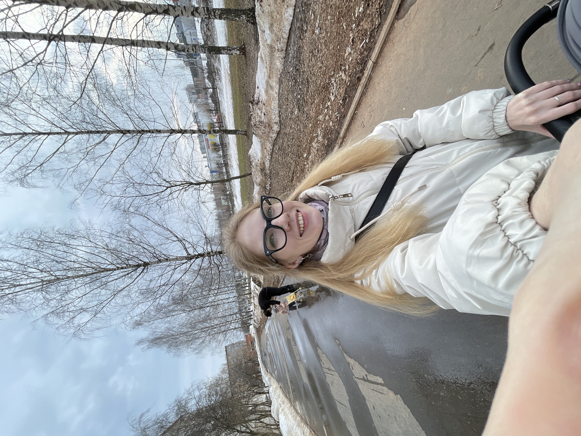 A woman with long blonde hair and glasses takes a selfie outside on a wet, paved path. She is wearing a white puffer jacket. Behind her, a person in a black outfit bends over near a yellow object on the wet pavement. The background shows bare trees, patches of melting snow, and buildings. The city is Izhevsk, Russia.