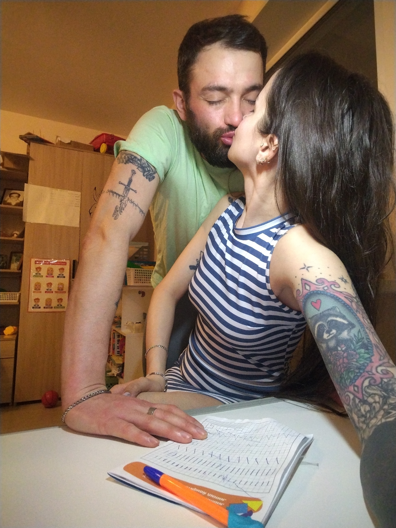 A man and a woman are indoors, possibly in an apartment, kissing. The man has a beard and is wearing a light green t-shirt. His left arm is visible, featuring a tattoo of a sword. The woman has long, dark hair and is wearing a striped blue and white top. Her right arm is covered with a detailed tattoo, including a raccoon design. They are positioned over a table on which a notebook is visible, filled with drawings, and an orange and blue marker. A bookshelf filled with various items and photographs is in the background. The location is Noviy, Russia.