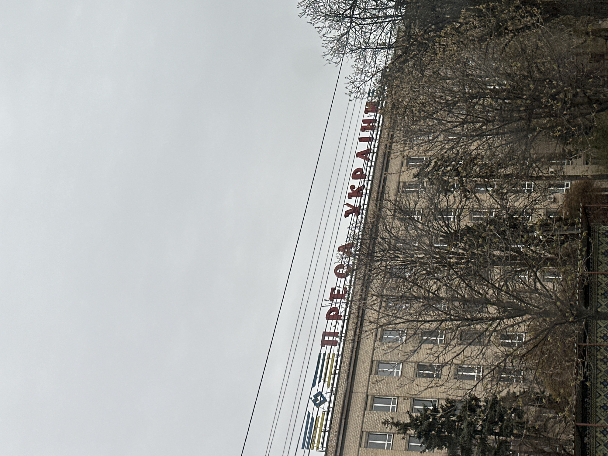 A low-angle shot of the "Press of Ukraine" building in Kyiv. The building is constructed of light-colored brick and has multiple windows on each level. The red Cyrillic text "ПРЕСА УКРАЇНИ" is prominently displayed on the building's facade. Power lines run diagonally across the frame, intersecting with the building and the sign. Bare branches of trees partially obscure the building. The sky is overcast and grey.