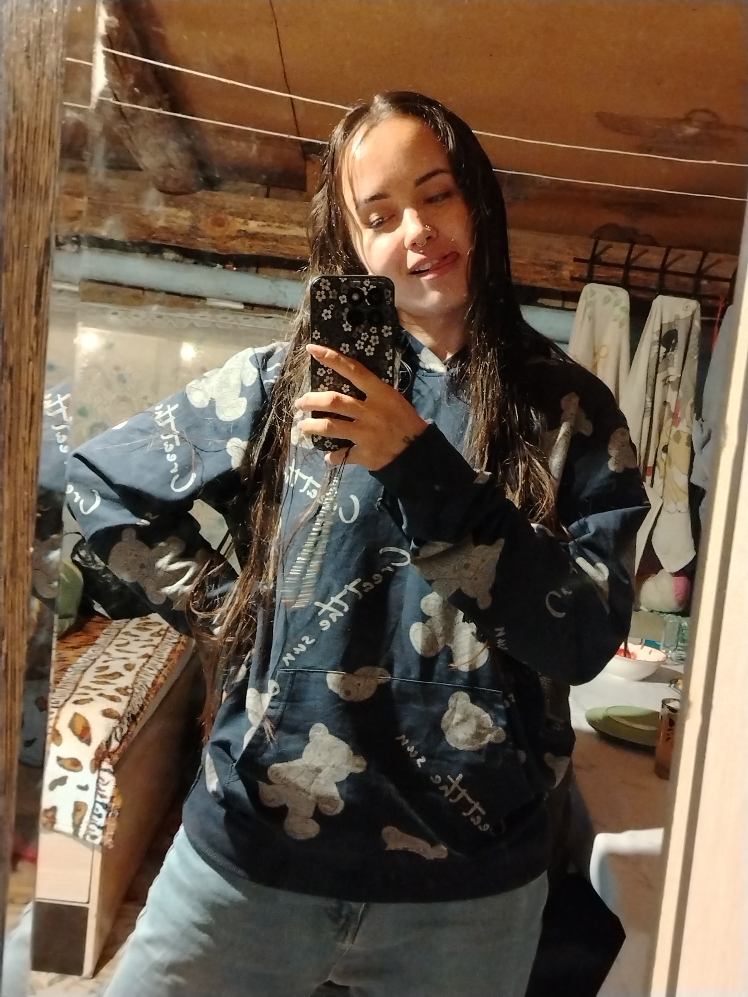 A young woman with long, dark hair and a nose piercing is taking a selfie in a mirror. She is wearing a dark blue hoodie with light-colored bear patterns and light blue jeans. The phone she is holding has a dark case with a floral design. She has her tongue slightly sticking out. The mirror reflects a room with a wooden ceiling and various objects in the background, including clothes hanging on a rack, a patterned rug on a bed, and items on a table. The mirror frame is made of wood. There is no information indicating this photo was taken in Гожан, Russia.