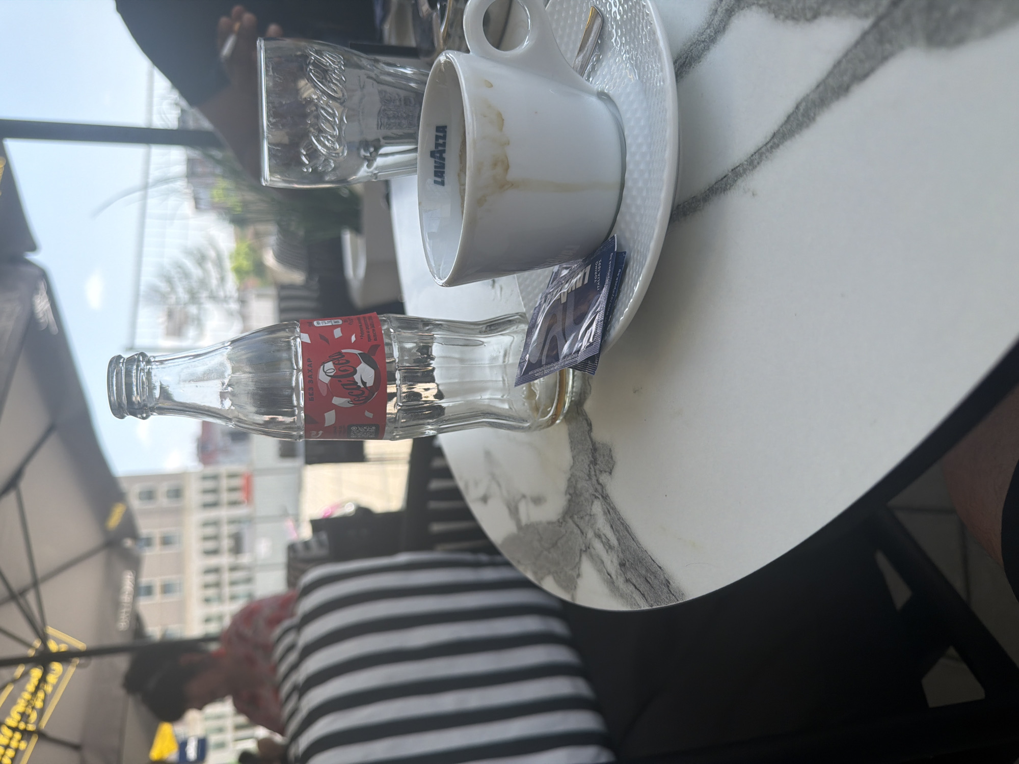 The image, rotated 90 degrees clockwise, presents a close-up view of a table at an outdoor cafe, likely during the daytime. The focus is on beverages consumed in an urban setting in Shumen, Bulgaria.
On a round table with a white, grey-veined marble pattern, several items are visible. An empty, clear glass Coca-Cola bottle lies horizontally, its red label prominently displaying "Coca-Cola" and the Cyrillic text "БЕЗ ЗАХАР" (sugar-free), along with a stylized red emblem. Adjacent to it is a white ceramic coffee cup, with "LAVAZZA" written vertically on its side, showing visible coffee stains inside. The cup sits on a white saucer with a textured, possibly dimpled, pattern, which also holds two small, purplish packets, likely sugar or sweetener, one bearing the "LAVAZZA TORINO ITALIANA 1895" branding. A small silver spoon rests on the saucer. Further back on the table, another clear glass with the "Coca-Cola" logo embossed on its surface is visible.
Two individuals are partially seen. In the lower left, a blurry person is seated, wearing a light-colored, possibly floral, shirt and appears to be leaning forward. A chair or cushion with prominent black and white horizontal stripes is also visible in this area. Another person's arm and dark clothing are partially visible in the upper left corner, resting on the table.
The background shows a blurred urban landscape with buildings under a bright sky, suggesting good weather. Part of a large, yellow-patterned outdoor umbrella is visible in the lower left, indicating shade at the cafe. The Cyrillic text on the Coca-Cola bottle is a notable detail, consistent with the specified location of Shumen, Bulgaria. The scene depicts a quiet moment at a cafe, following the consumption of drinks.