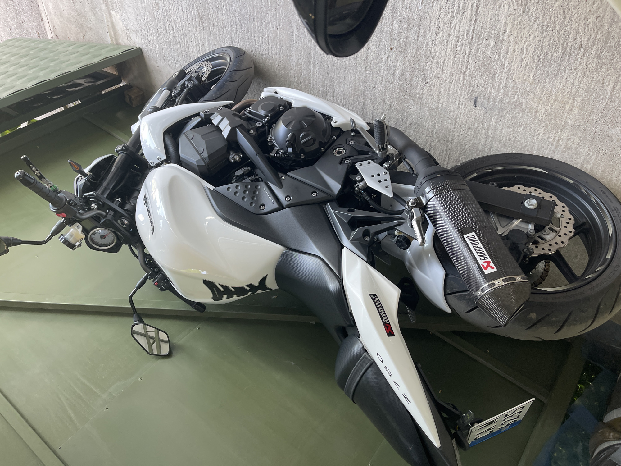 The image shows a white and black Kawasaki Z750 motorcycle parked in what appears to be a garage or storage area. The motorcycle is positioned at an angle on a green, possibly metallic surface. The exhaust system features an Akrapovic muffler. A side mirror and a license plate are visible. The background includes a concrete wall and a partial view of a structure with a green roof. The location is not discernable to Sofia, Bulgaria.