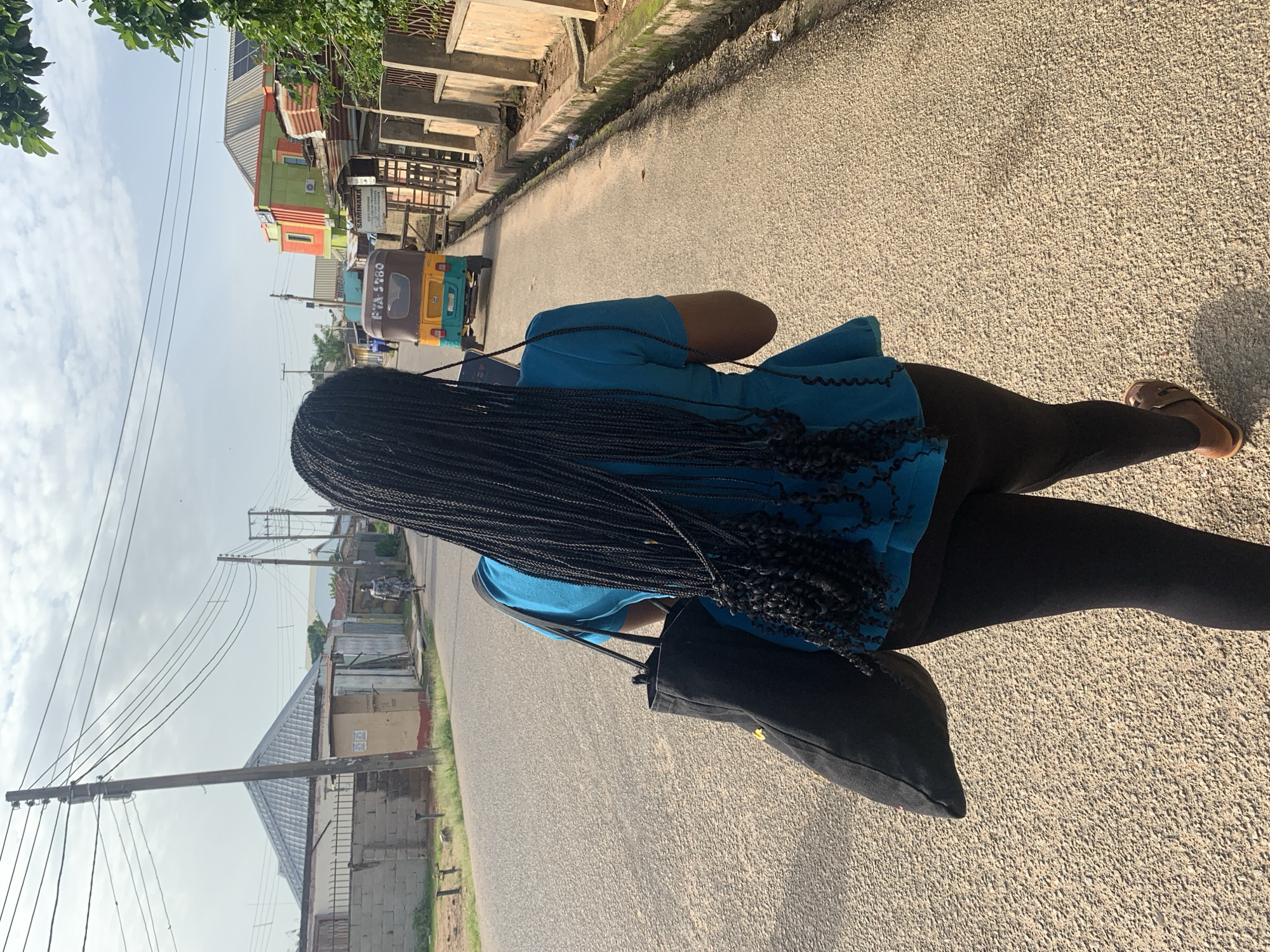 The image shows a woman from the back, walking down a street in Jos, Nigeria. She has long, dark braided hair and is wearing a blue top and black leggings. She is carrying a black bag over her shoulder. The street is paved and lined with buildings on either side. There is a small, colorful auto-rickshaw driving in the distance. The sky is overcast, suggesting it might be a cloudy day. The scene appears to be daytime. The woman's posture suggests she is walking with purpose. There is a sign visible on one of the buildings with the text "SAMINAKA."