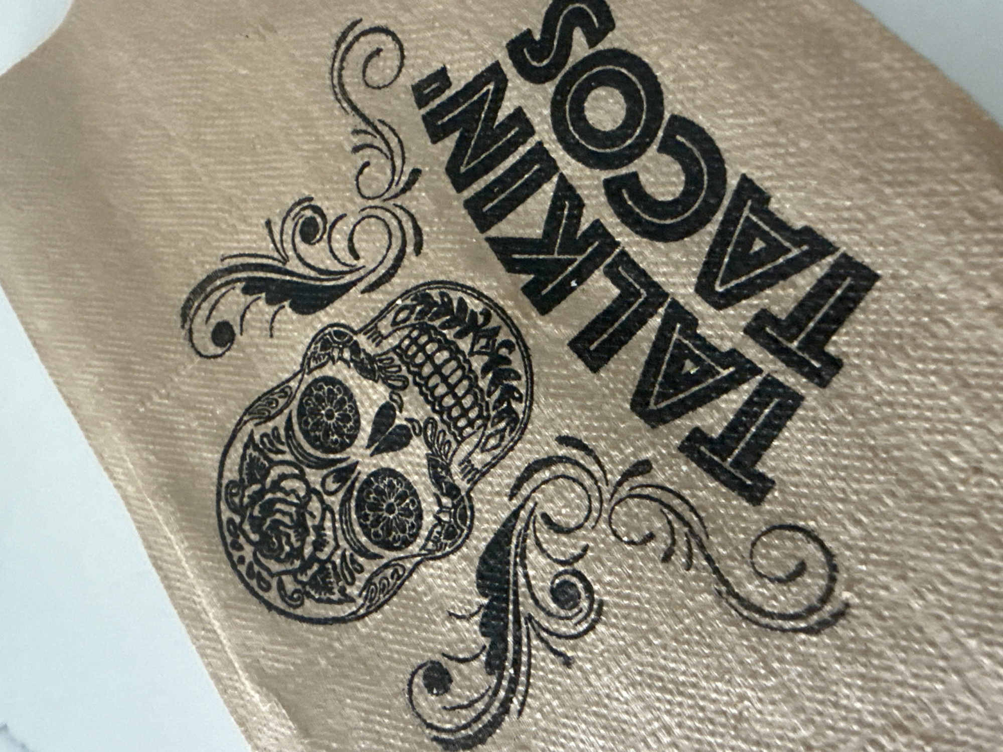 The image shows a tan-colored napkin with a sugar skull design and the words "TALKIN' TACOS" printed in black ink. Ornate black swirls also decorate the napkin. The exact location within Davie, United States of America is not discernable from the image.