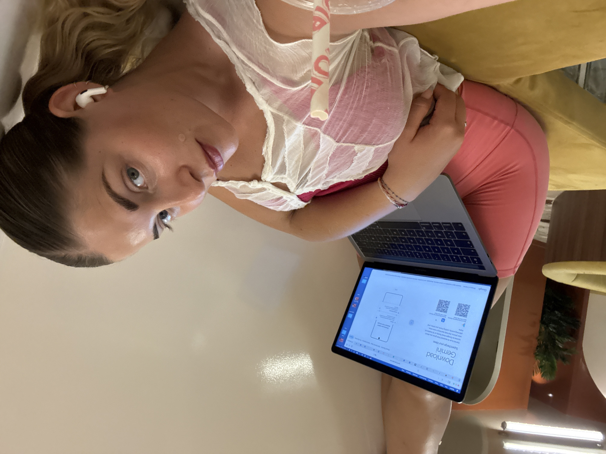 A person is lying down, propped up on a yellow couch, and using a laptop. The person has blonde hair, light skin, and is wearing a sheer white top over a pink bra and pink leggings. They are also wearing white earbuds. The laptop screen displays information about downloading "Gemini" and has two QR codes. The image is taken indoors, with soft lighting suggesting it could be daytime. The person's gaze is directed towards the laptop, with a neutral expression. The background is mostly out of focus but includes part of the yellow couch and a hint of an indoor plant. The scene appears relaxed and casual.