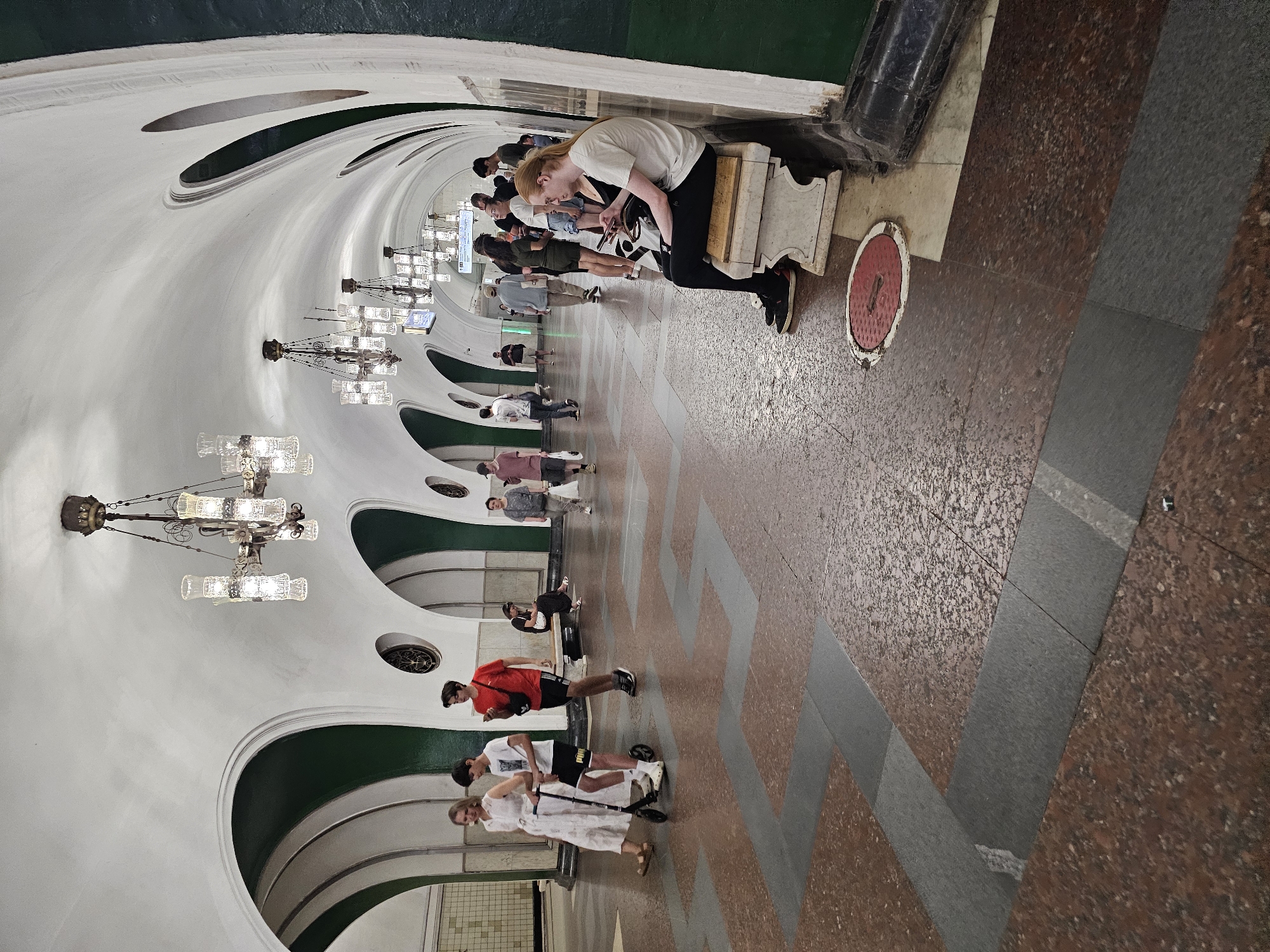 This image depicts the interior of a Moscow Metro station, likely a deep underground station given the ornate architecture. The station features a long, vaulted ceiling with decorative chandeliers, and arched openings revealing green tiled walls. The floor is a patterned mosaic of red and grey granite tiles.
In the foreground, a woman in a white dress walks alongside a young boy on a scooter. To their right, a young man in a red t-shirt stands looking at his phone. Further down the platform, several other individuals are visible, some standing and some sitting on a bench. A woman with blonde hair and a white t-shirt is seated on the bench, also looking at her phone. The overall atmosphere is one of a busy public transport hub.