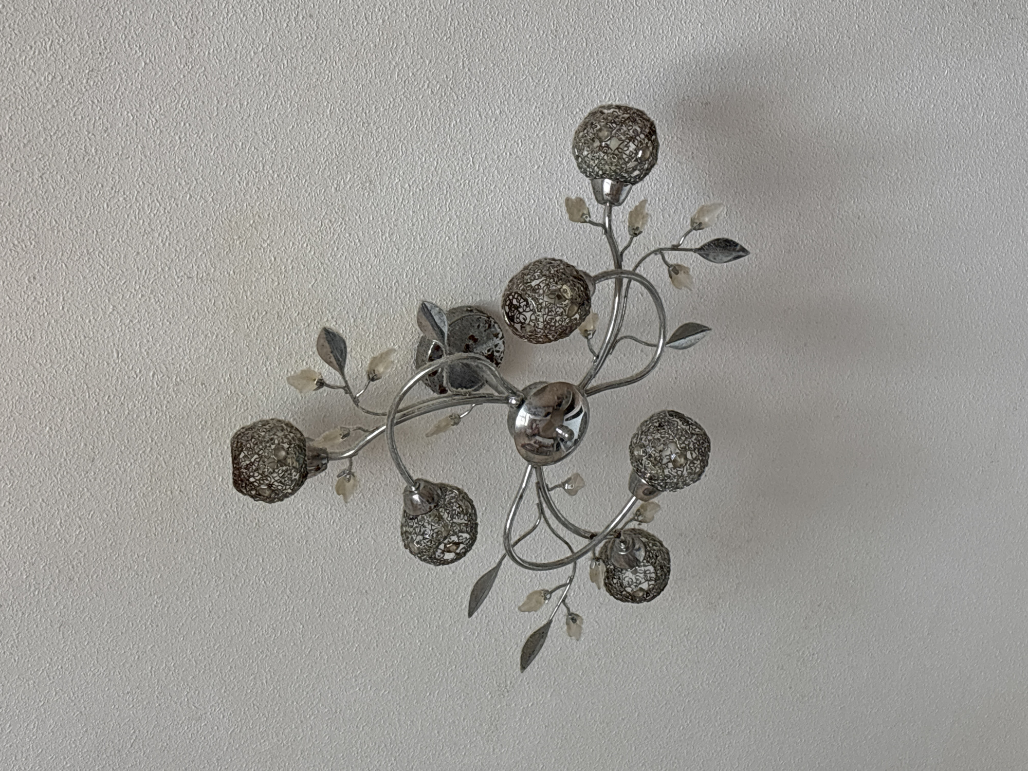 This image features a close-up, overhead view of a decorative light fixture mounted on a textured, off-white ceiling. The fixture is metallic, likely silver or chrome, and designed in an organic, branch-like style. Several spherical lampshades, constructed from an intricate, mesh-like metal, are distributed along the branches, resembling glowing orbs. Interspersed with the lampshades are small, translucent decorative elements, possibly made of glass or plastic, shaped like buds or leaves, and some metallic leaf-shaped accents. A central, polished metallic sphere reflects the surroundings. The lighting fixture appears to be a decorative ceiling lamp, possibly for an interior living space. No people are visible, and there are no strong cues about the time of day or weather, other than the typical indoor lighting implied by the presence of a lamp. The location context provided is Orihuela, Spain, which is a general geographical identifier and does not offer specific visual clues within the image itself. There is no visible text in the image.