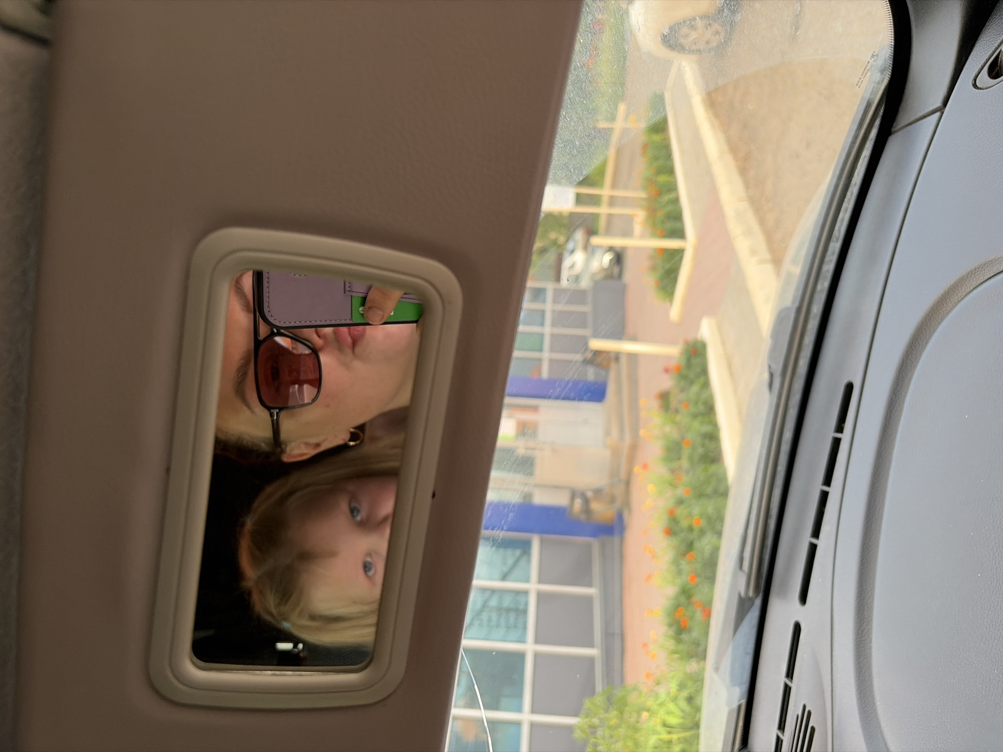 This media file shows a reflection in a car's visor mirror. In the mirror, a woman wearing sunglasses is visible, holding a phone with a purple case. Another reflection shows a child with blue eyes and blonde hair. The background in the reflection is blurry, showing buildings with blue and white accents, and some greenery with small orange flowers, suggesting an outdoor setting. The time of day appears to be daytime, with bright lighting. The overall scene suggests the two individuals are in a car, possibly parked. No visible text is present in the image.